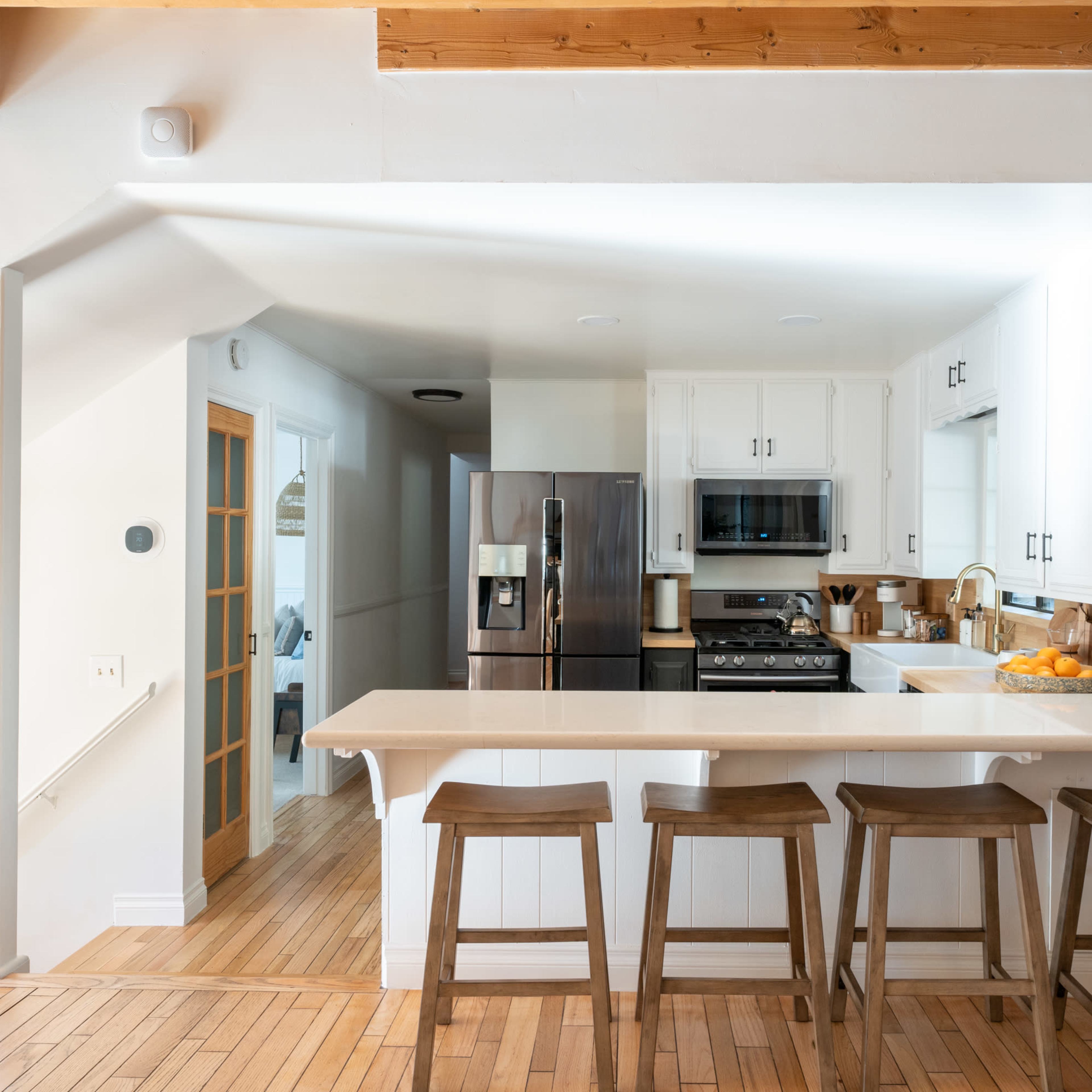 A bright kitchen features a countertop bar with four wooden stools, a stainless steel refrigerator, and a gas stove, all surrounded by white cabinetry and wood accents.