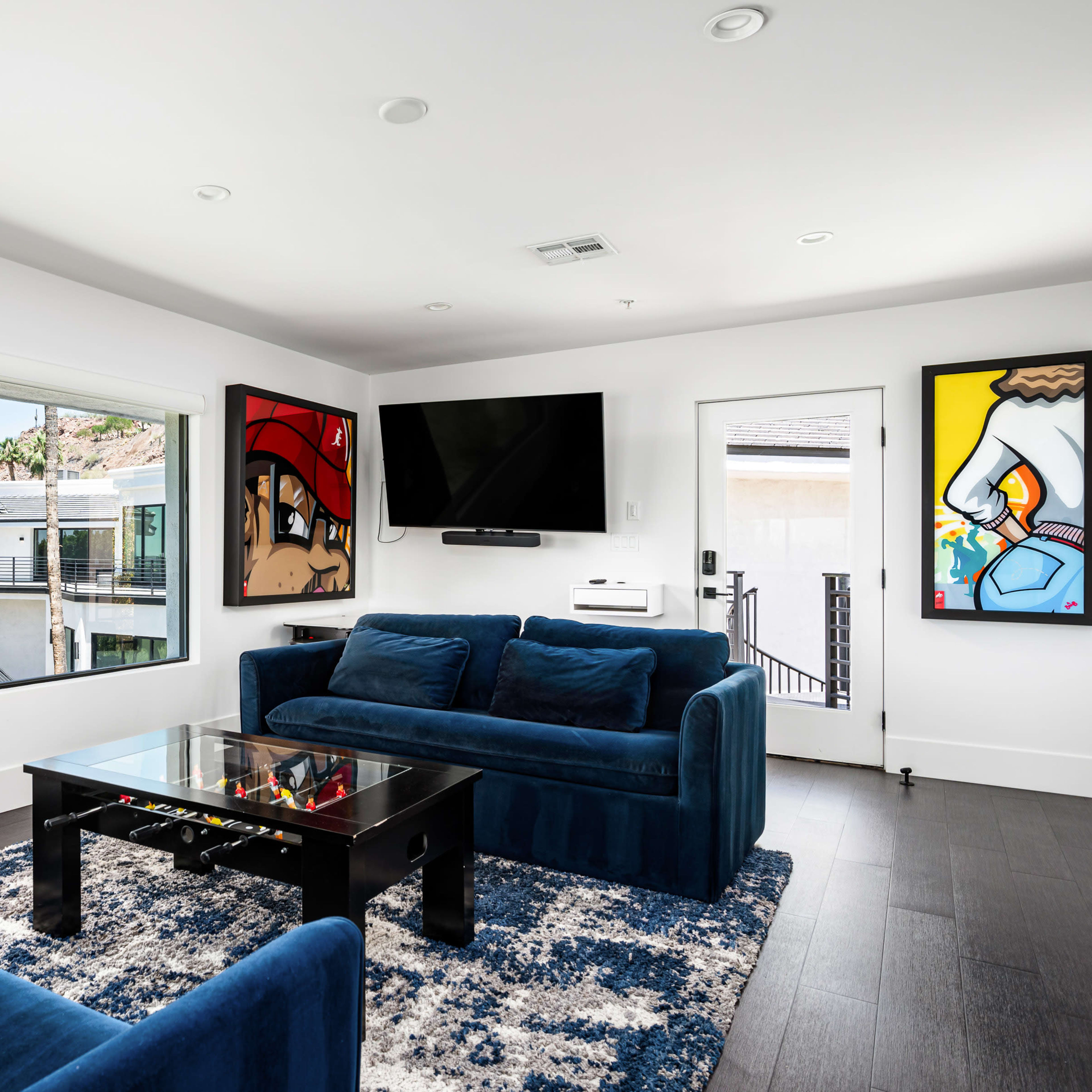 A modern living room features blue sofas and a black coffee table, with large windows providing a view of palm trees and outdoor space.