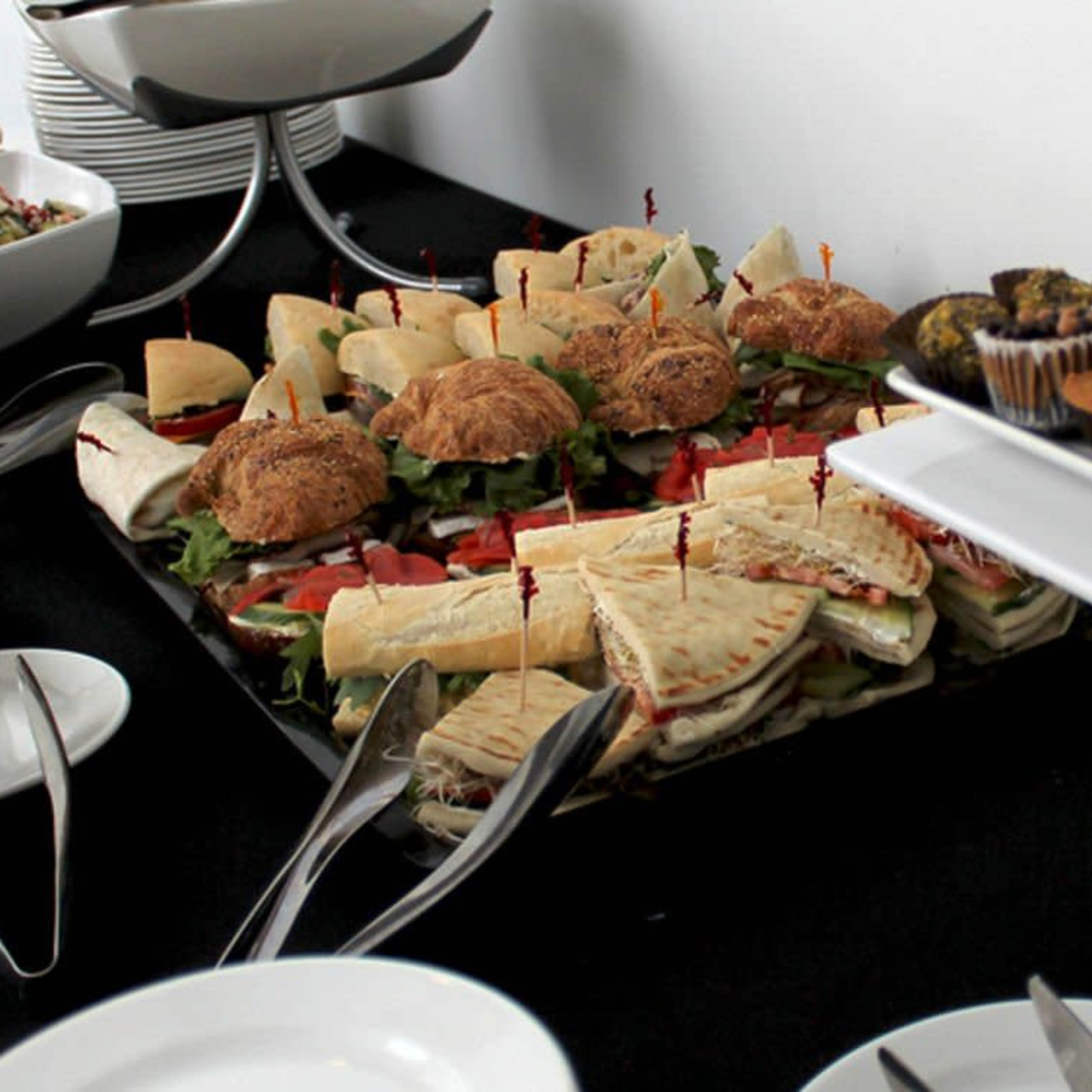 A catered food spread features sandwiches, salads, and an assortment of pastries on a black tablecloth.