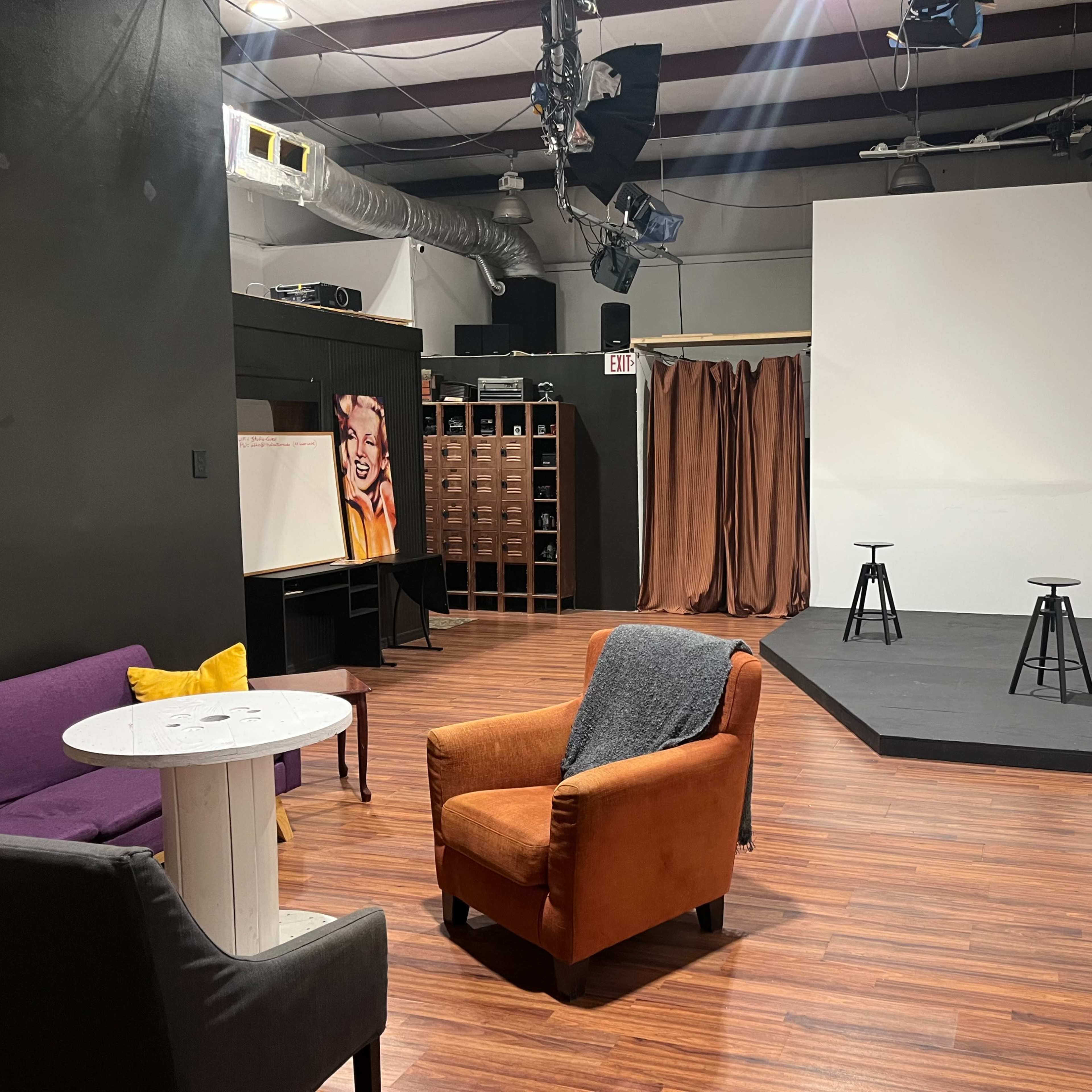 A sparse theater setup with a small stage, three black stools, a curtain, and seating arrangements including an orange armchair and a purple couch.