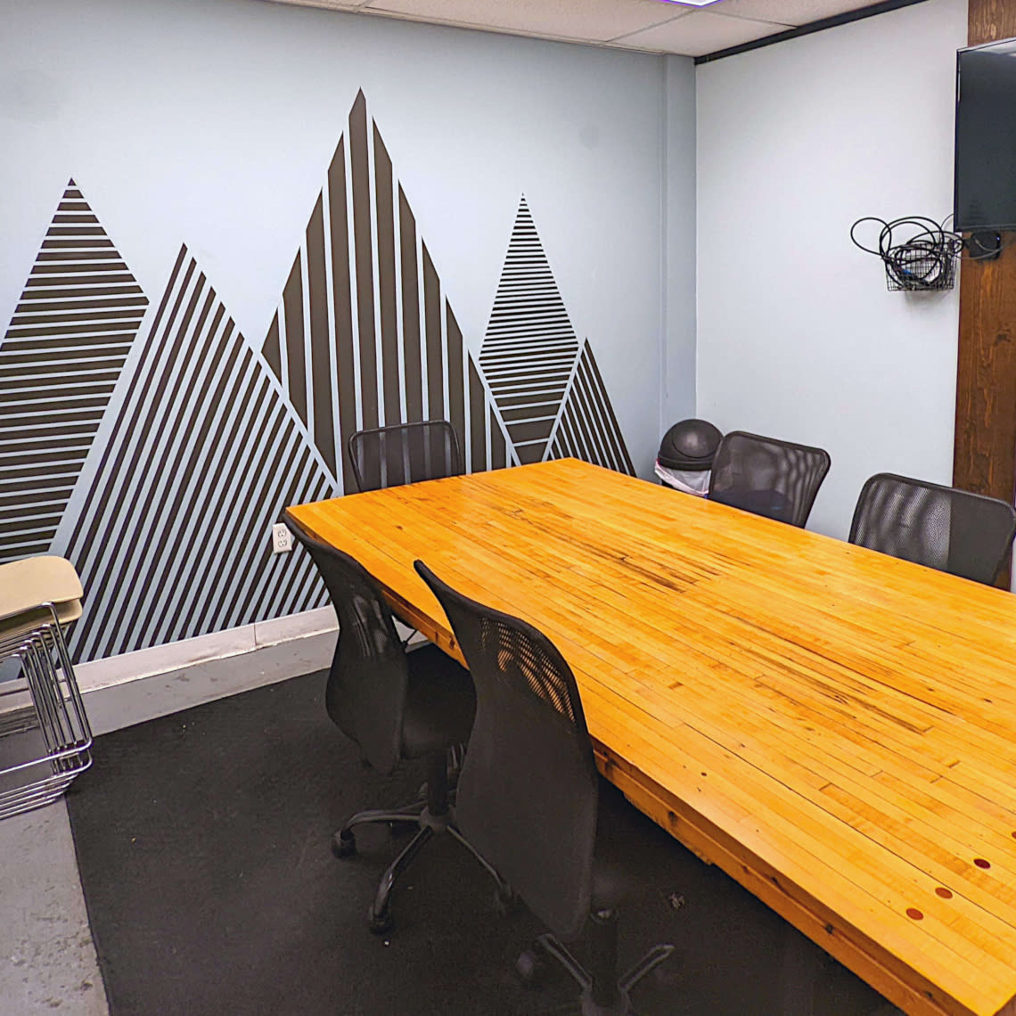 A conference room with a large wooden table surrounded by black chairs, a television mounted on the wall, and a geometric mountain mural on the opposite wall.