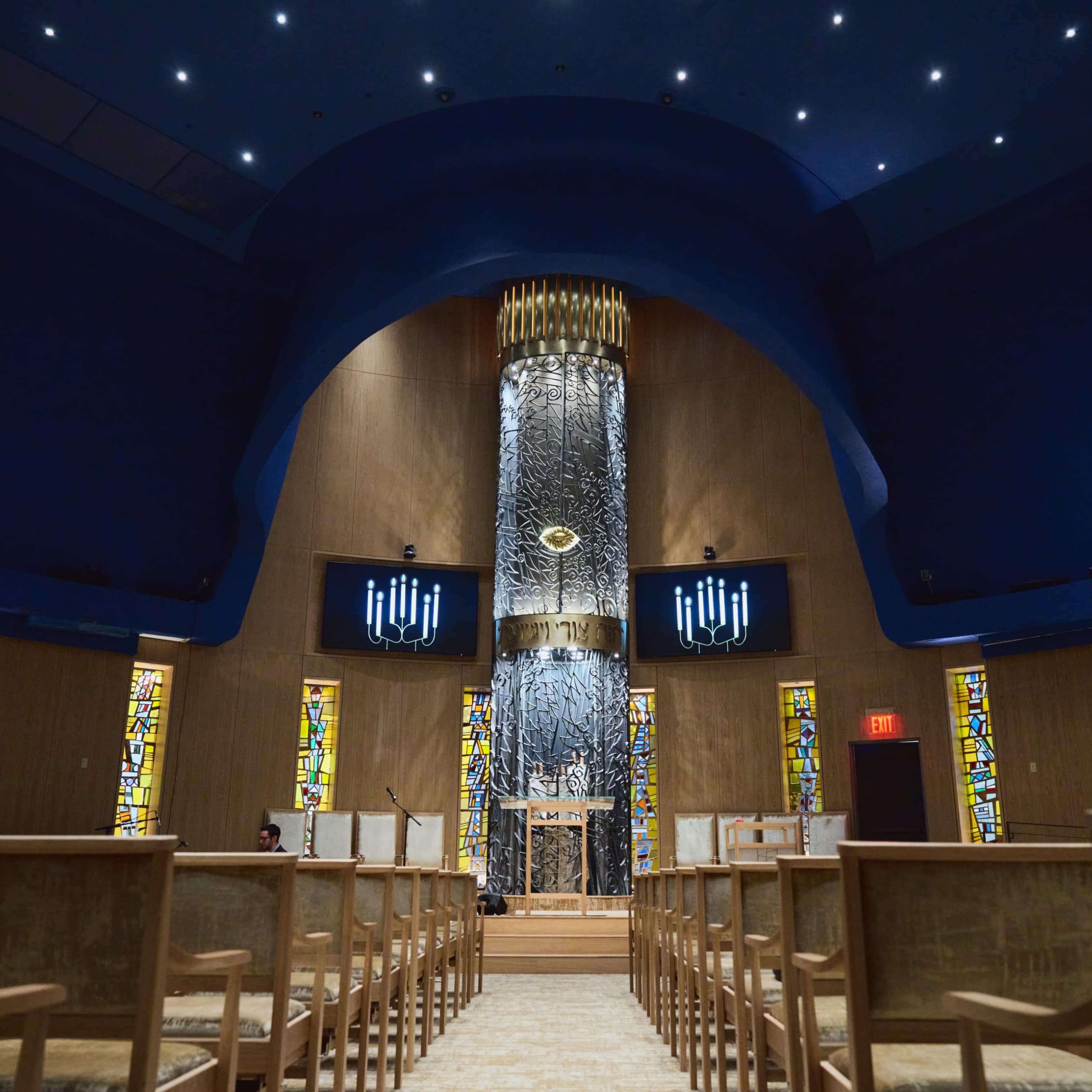 The interior of a synagogue features a central ornate ark surrounded by vibrant stained glass windows and rows of wooden chairs facing forward.