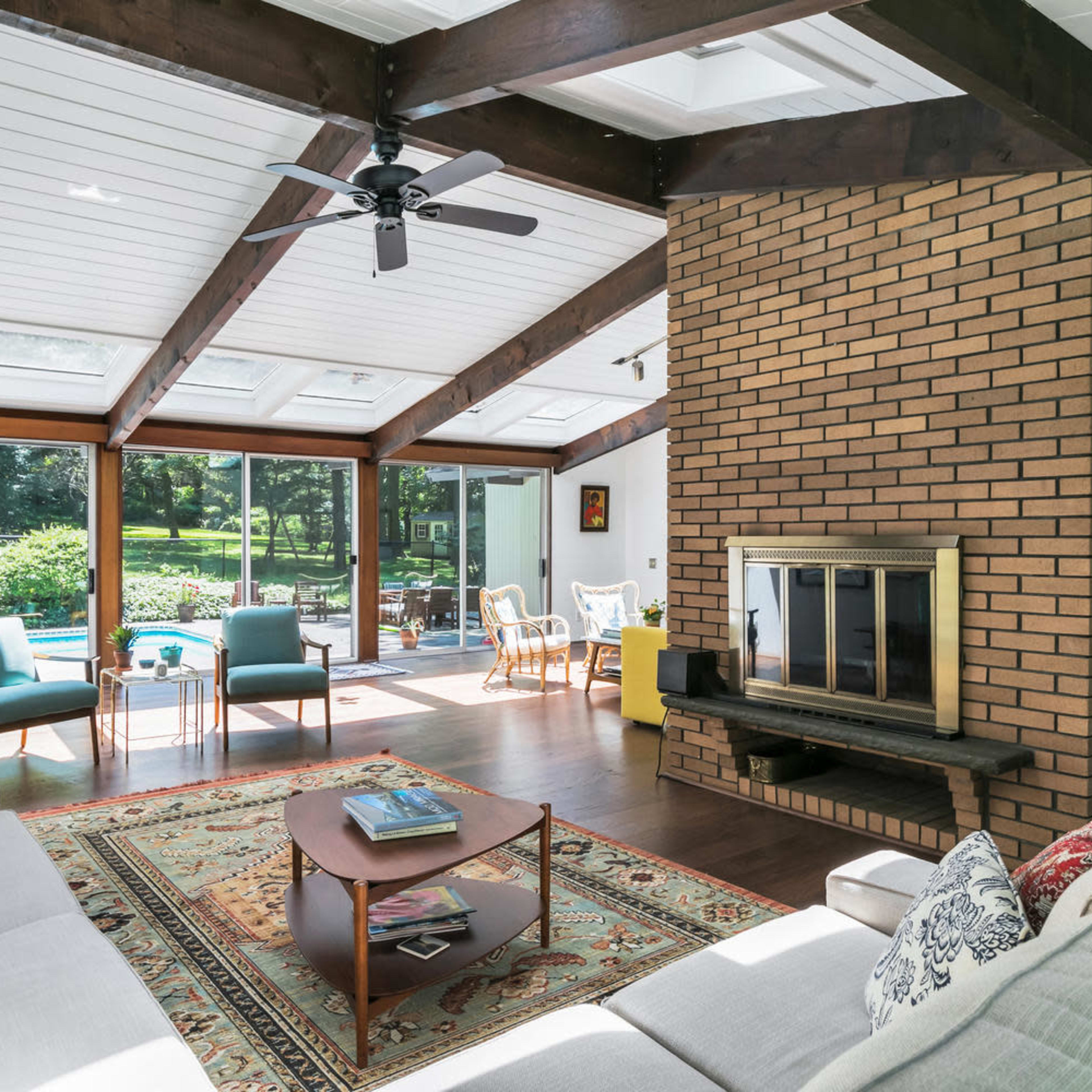A spacious living room features a large window wall overlooking a garden, a brick fireplace, and modern furniture arranged around a central coffee table.