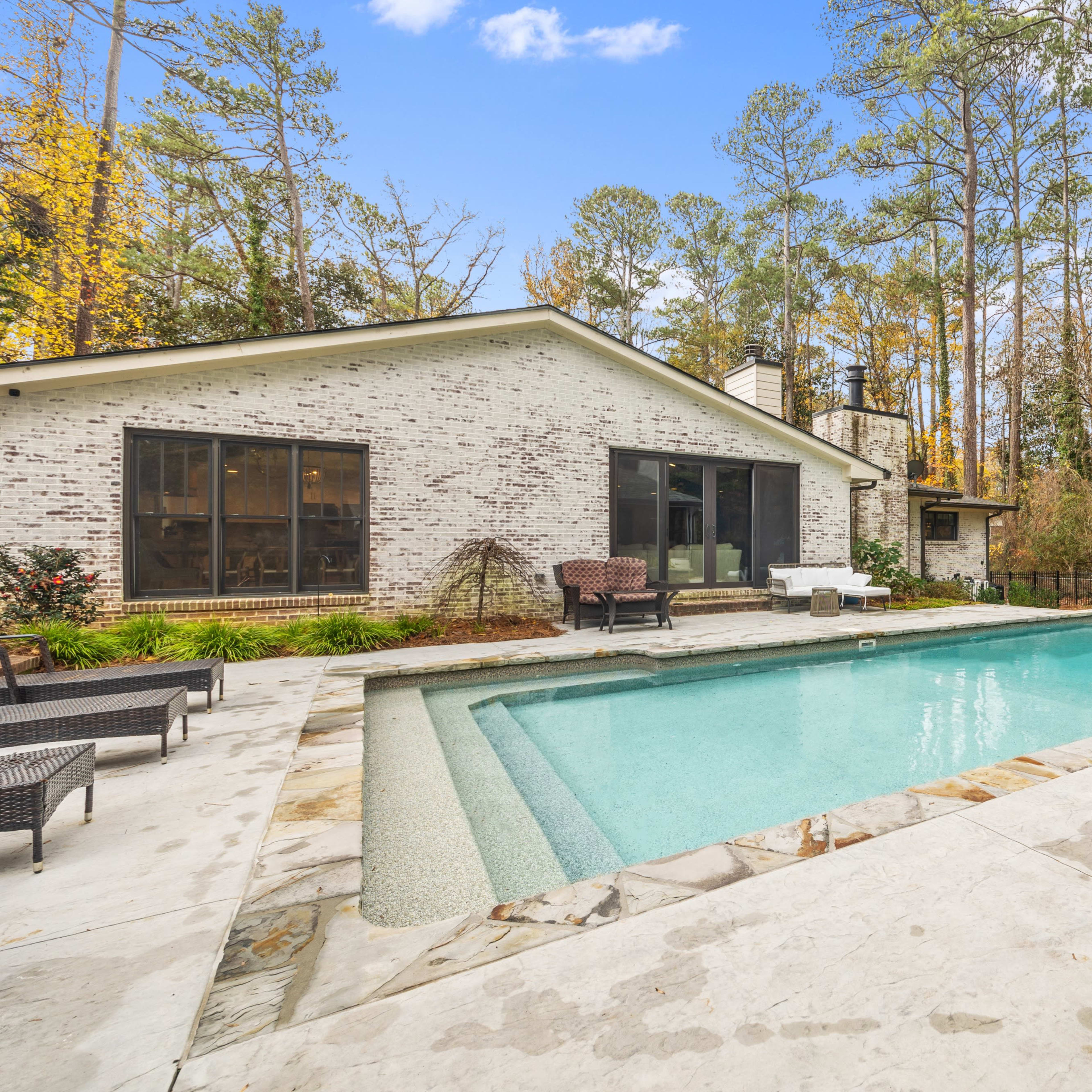 A backyard with a swimming pool surrounded by a stone patio and vegetation, adjacent to a house with large windows.