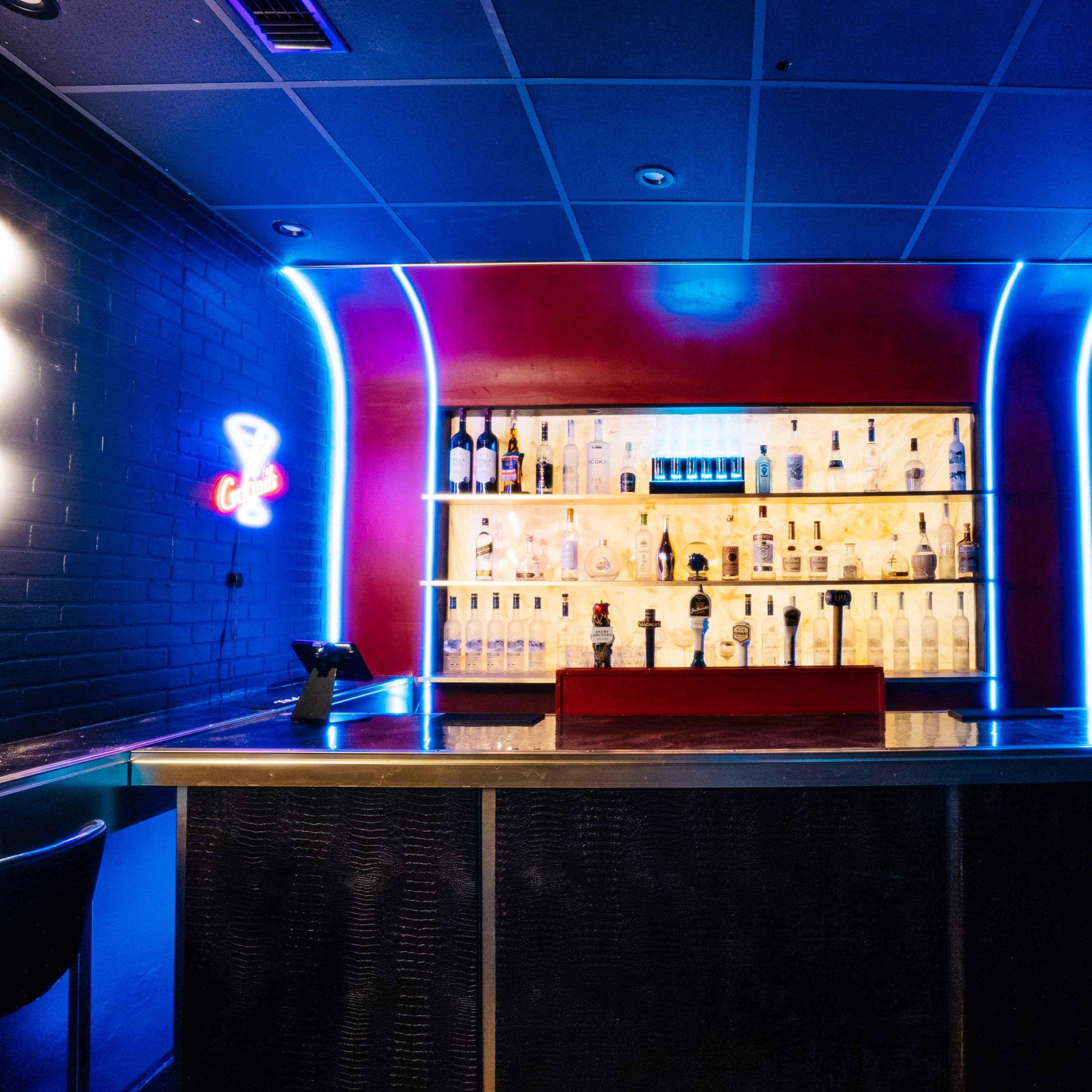 A bar with illuminated shelves displaying various bottles is set against a backdrop of red and black walls.