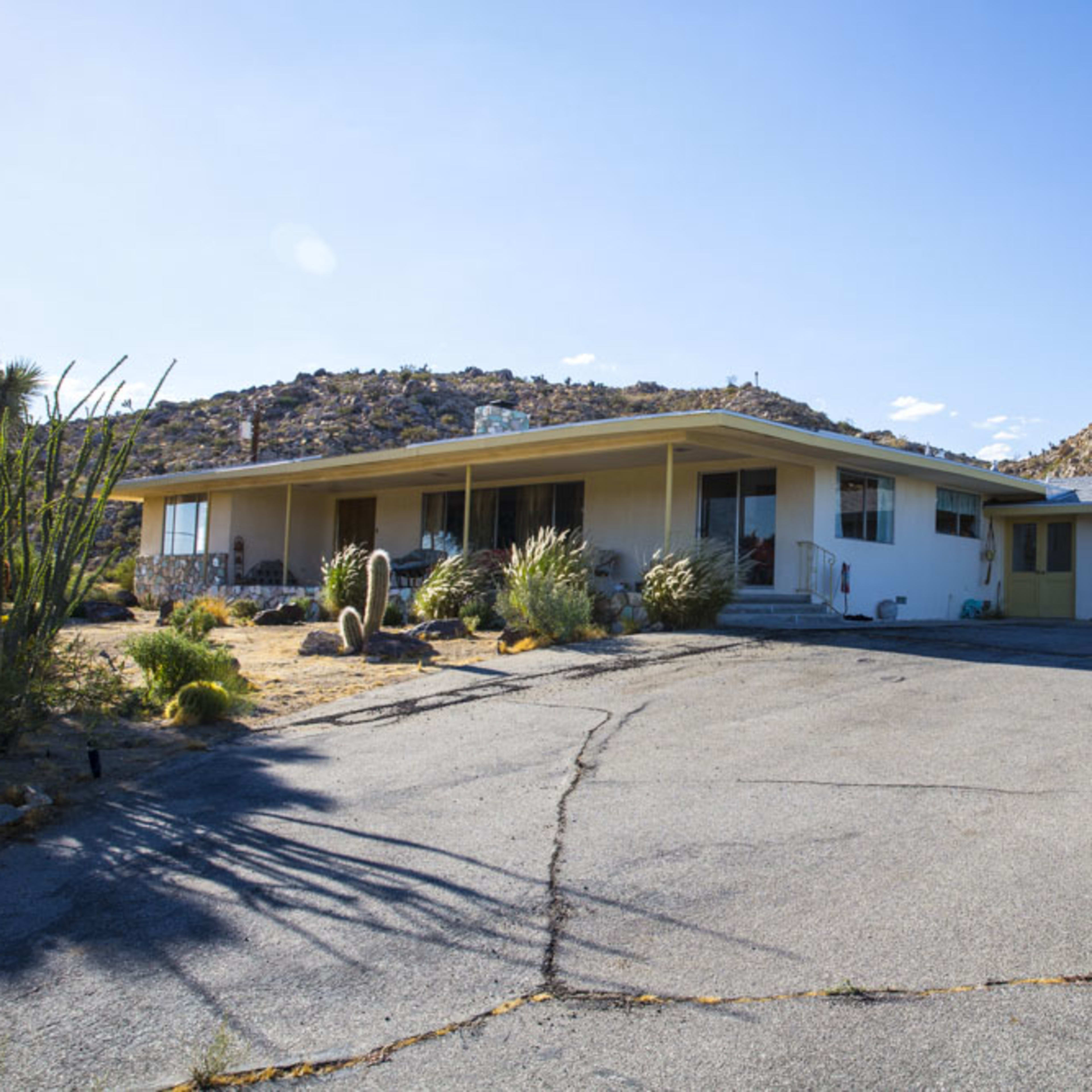 A single-story house with a flat roof is situated on a paved driveway against a backdrop of rocky terrain and desert vegetation.