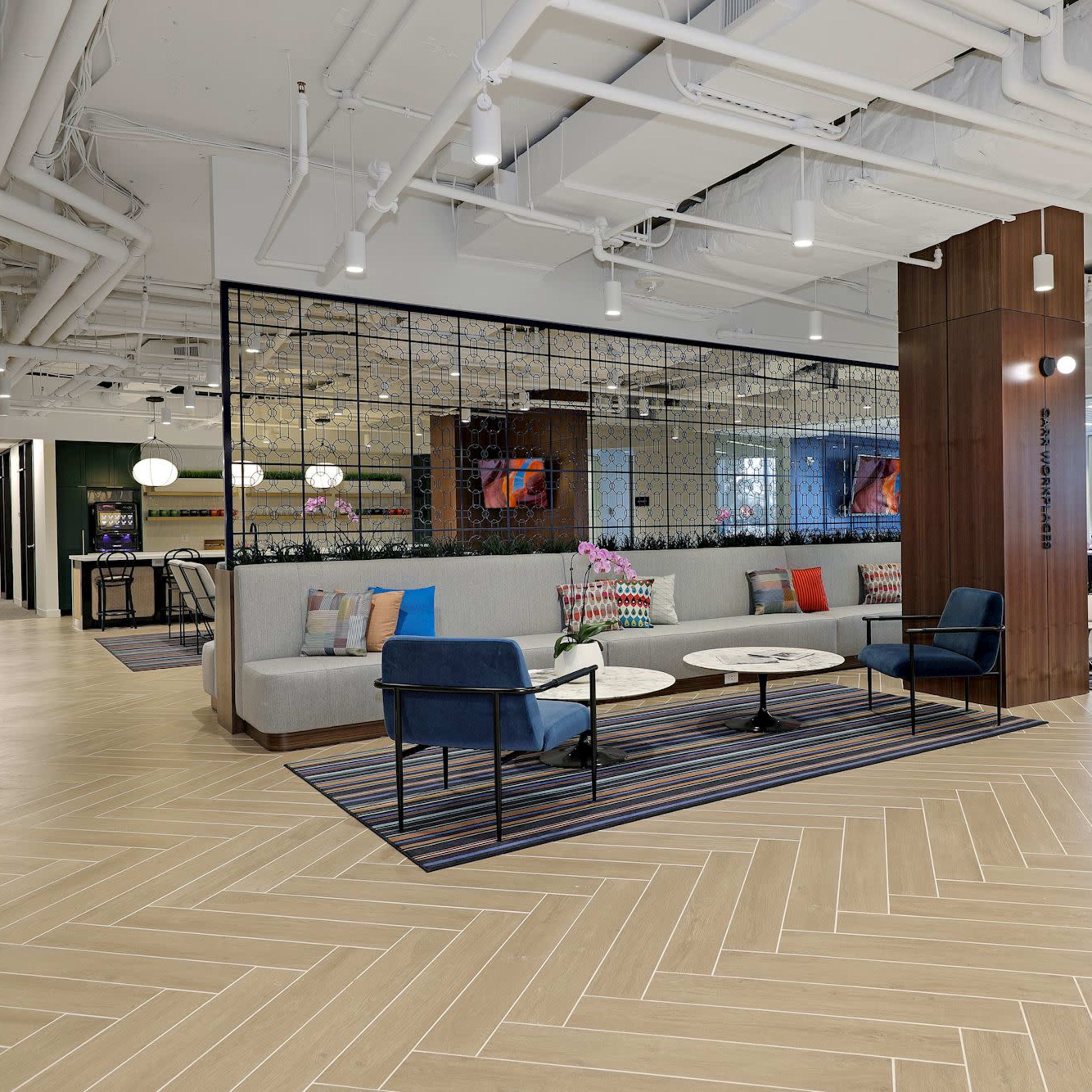 A modern office lounge area with comfortable seating, a glass partitioned meeting space, and wooden floors.