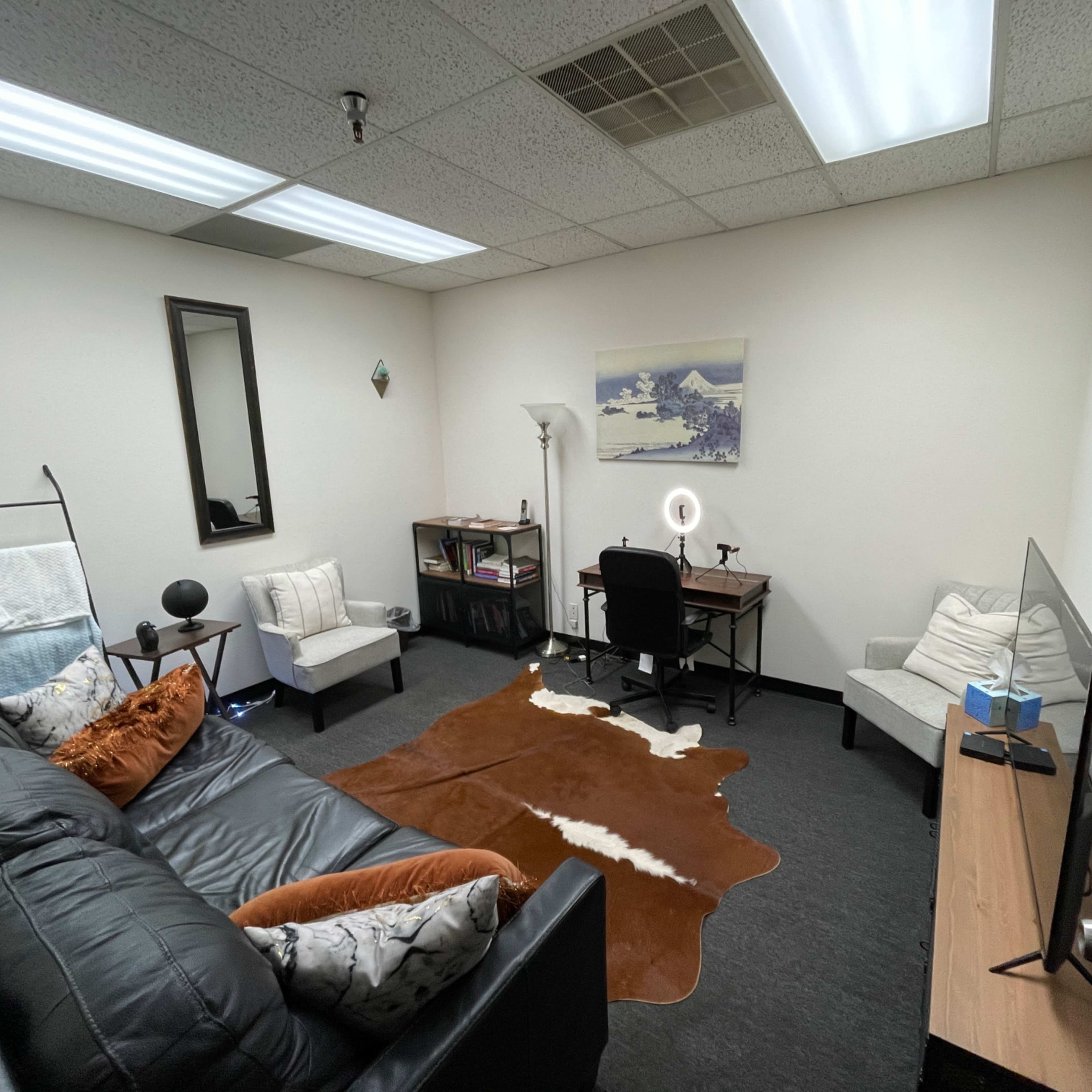 A small office space featuring a black couch, a desk with a chair, and decorative elements like a mirror and a painting on the wall.