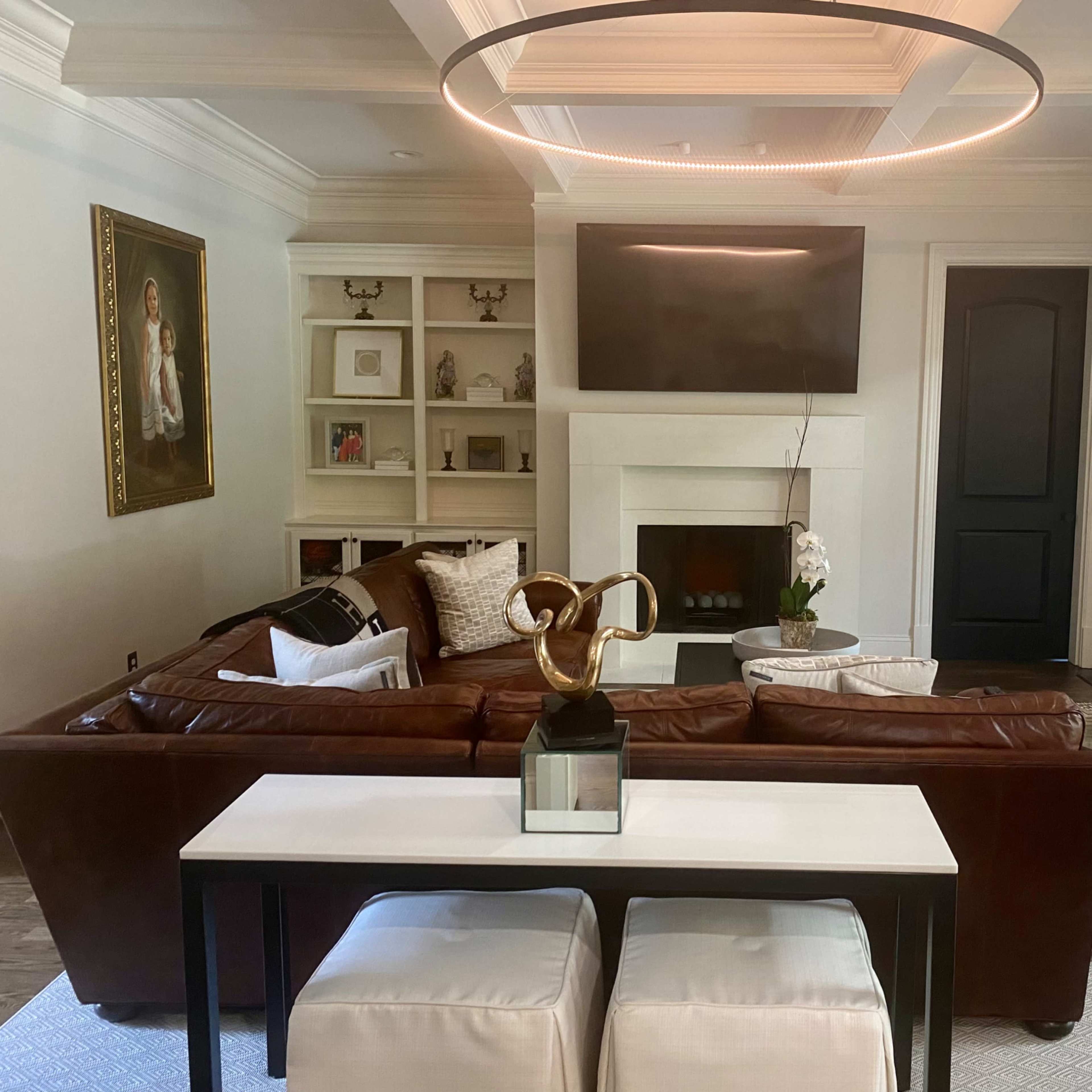A modern living room with a brown leather sectional sofa, a circular ceiling light, a mounted flat-screen TV above a fireplace, and large black-framed doors leading to an outdoor area.