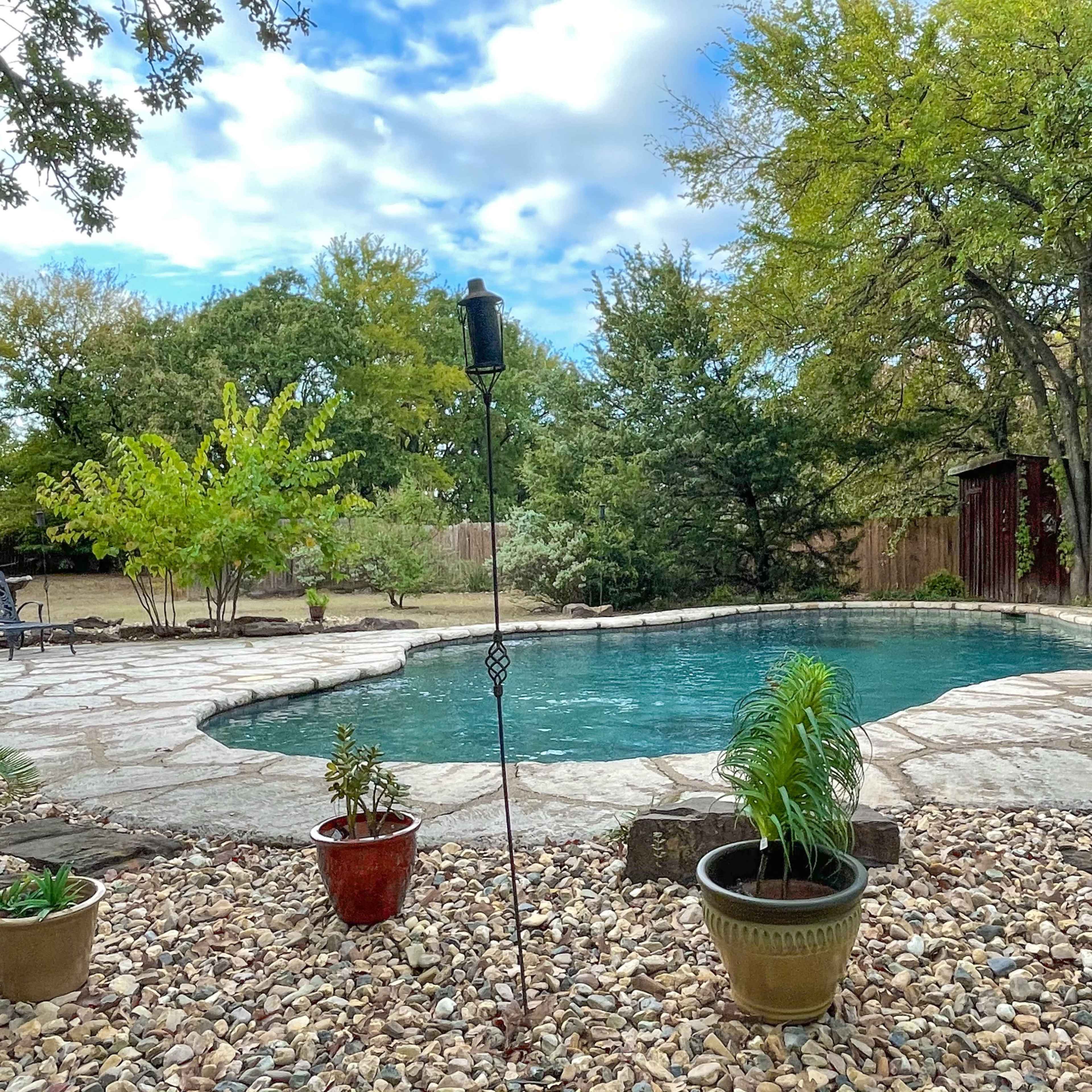 A landscaped backyard featuring a stone patio surrounding a clear swimming pool, with potted plants and trees in the background.