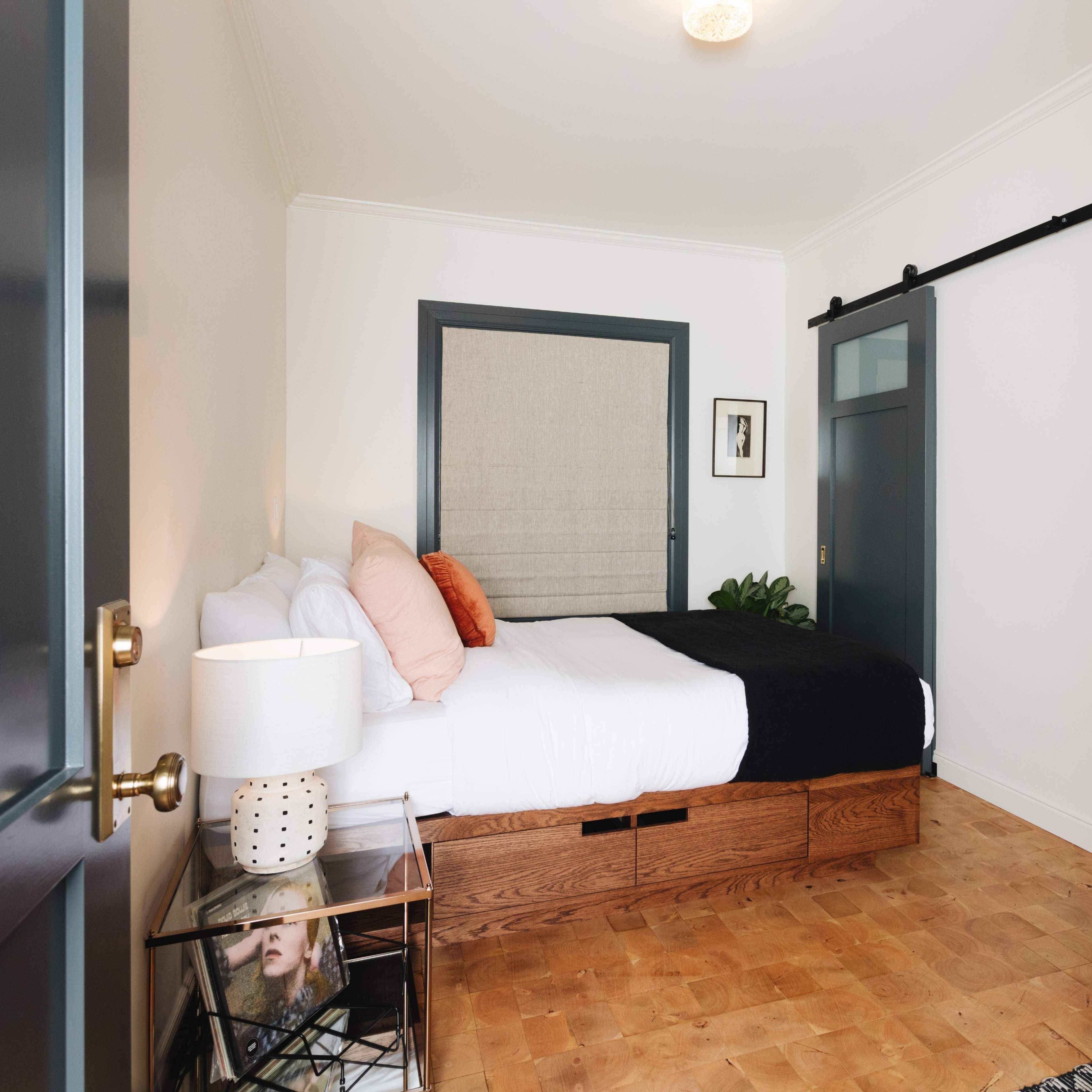 A modern bedroom features a bed with white linens and orange accents, a wooden storage base, and sliding barn door, along with a glass shower visible in the background.