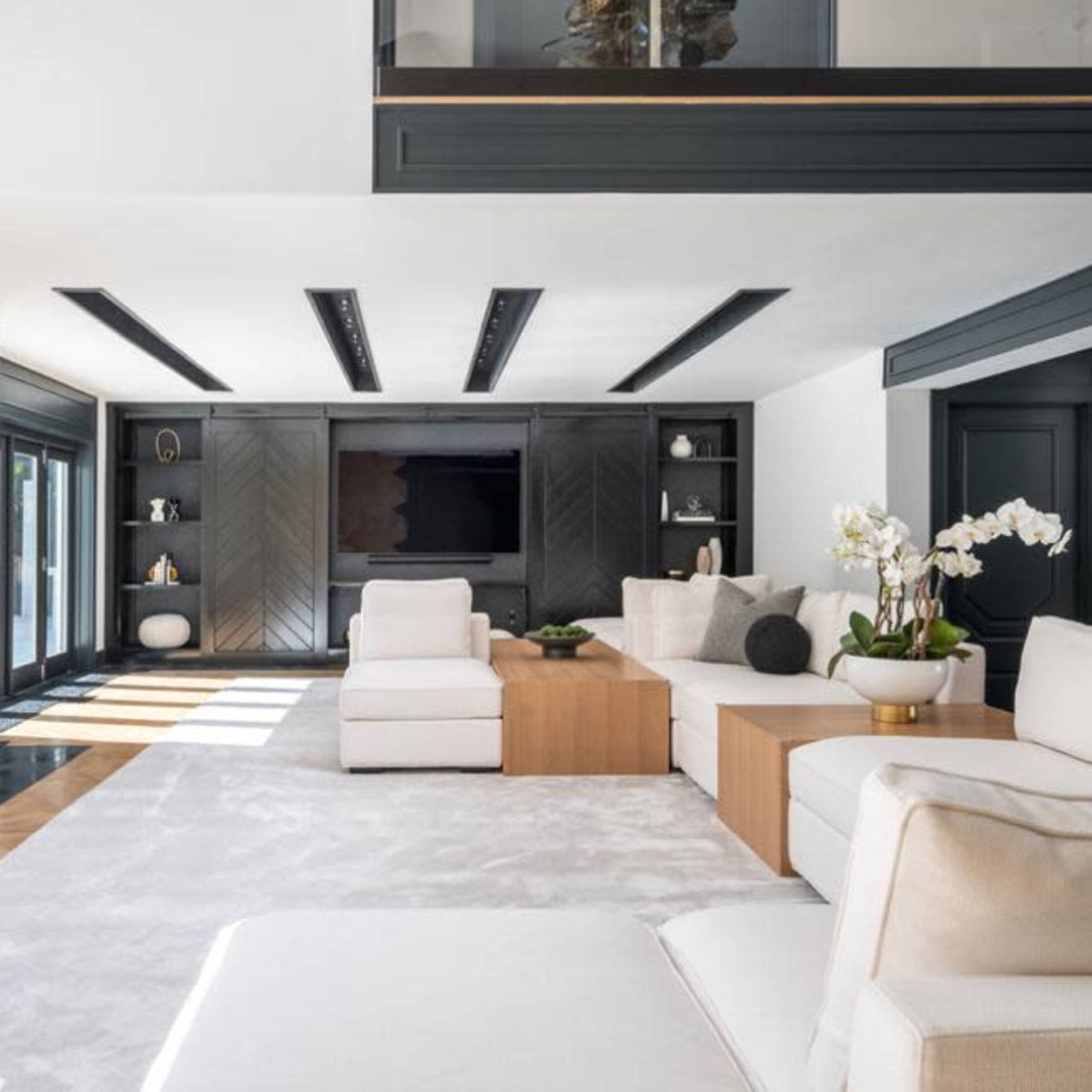 A spacious living room features a fireplace with a marble surround, light-colored sofas, and a wooden coffee table, all illuminated by natural light from large windows.