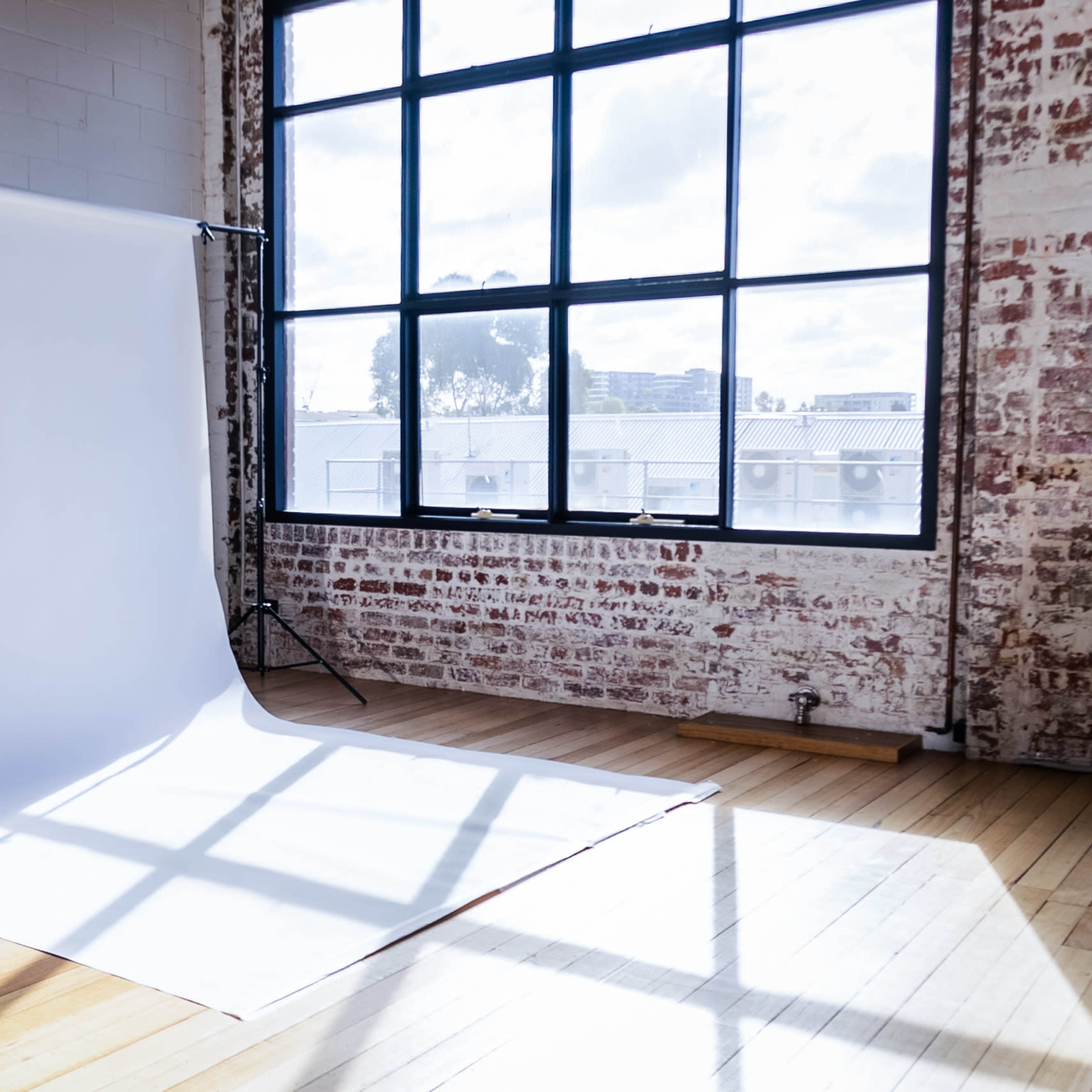 A spacious photography studio features a large white backdrop, positioned against a brick wall, with natural light streaming through a large window.