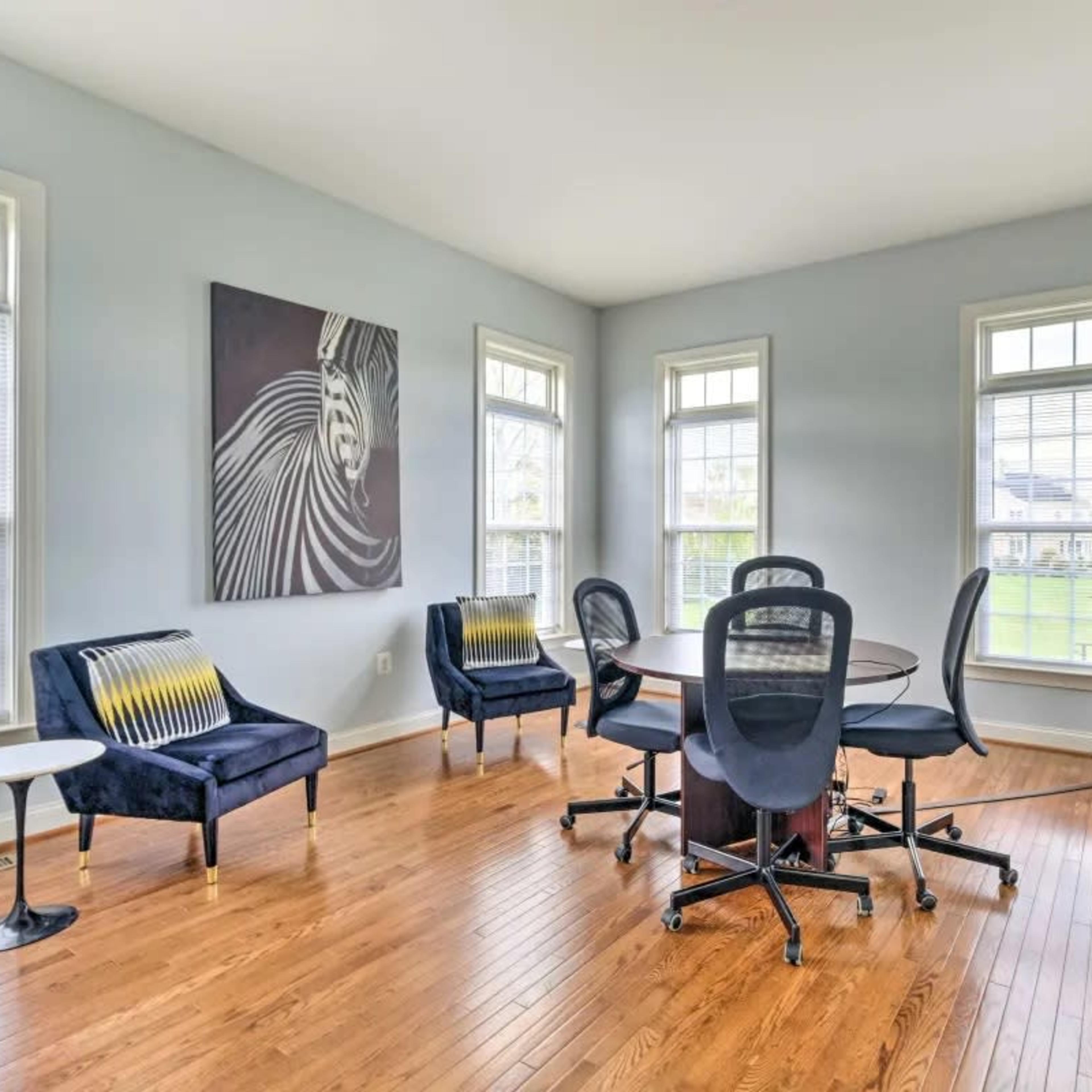 A bright, airy meeting room features a round table surrounded by six office chairs, with two blue armchairs and large windows allowing natural light to enter.