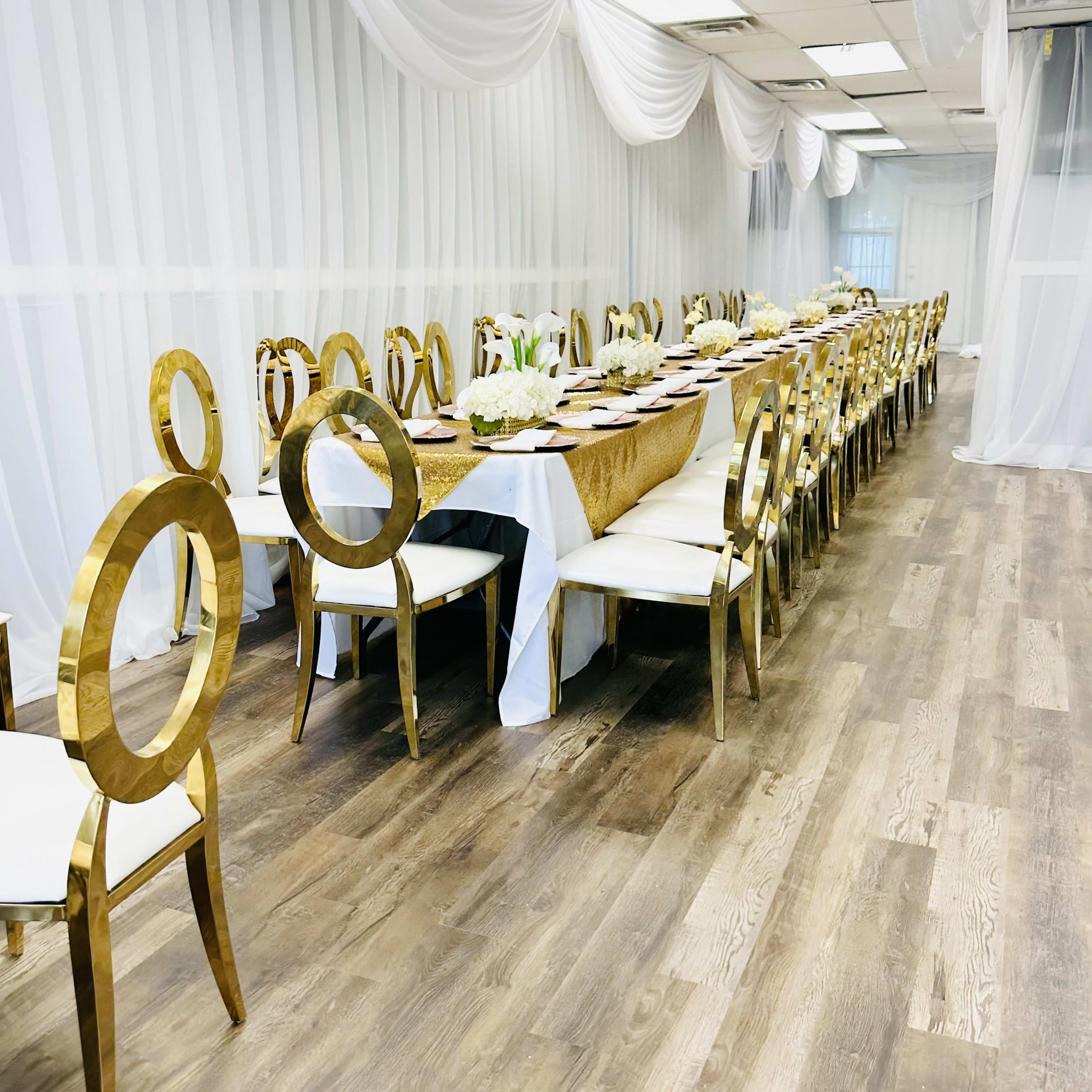 A long table is set with elegant decor and surrounded by gold chairs in a brightly lit room with white drapery.
