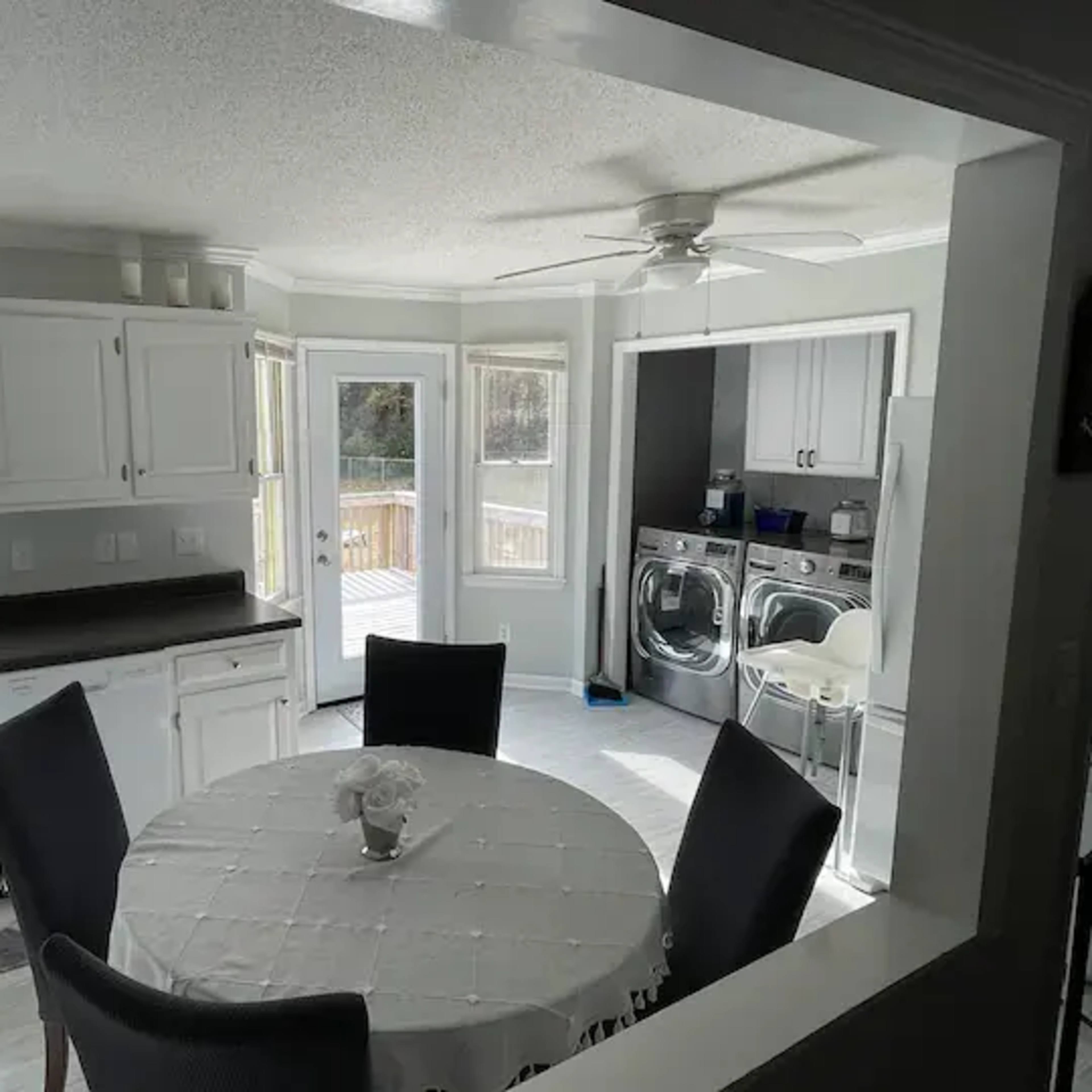 A bright kitchen and laundry area with a circular dining table in the foreground, white cabinets, and large windows.