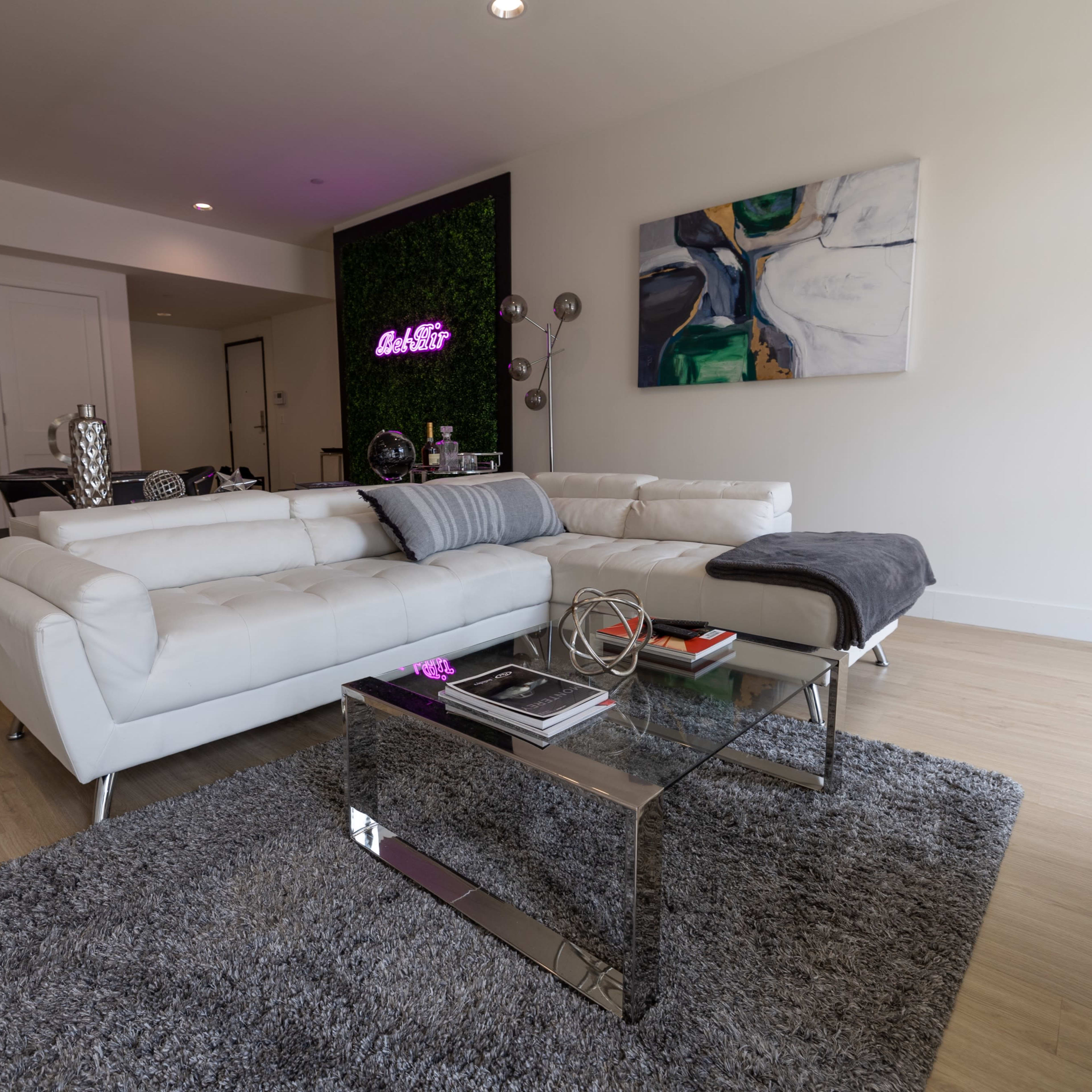 A modern living room featuring a white sectional sofa, a glass coffee table, and a decorative wall with vibrant artwork and a neon sign.