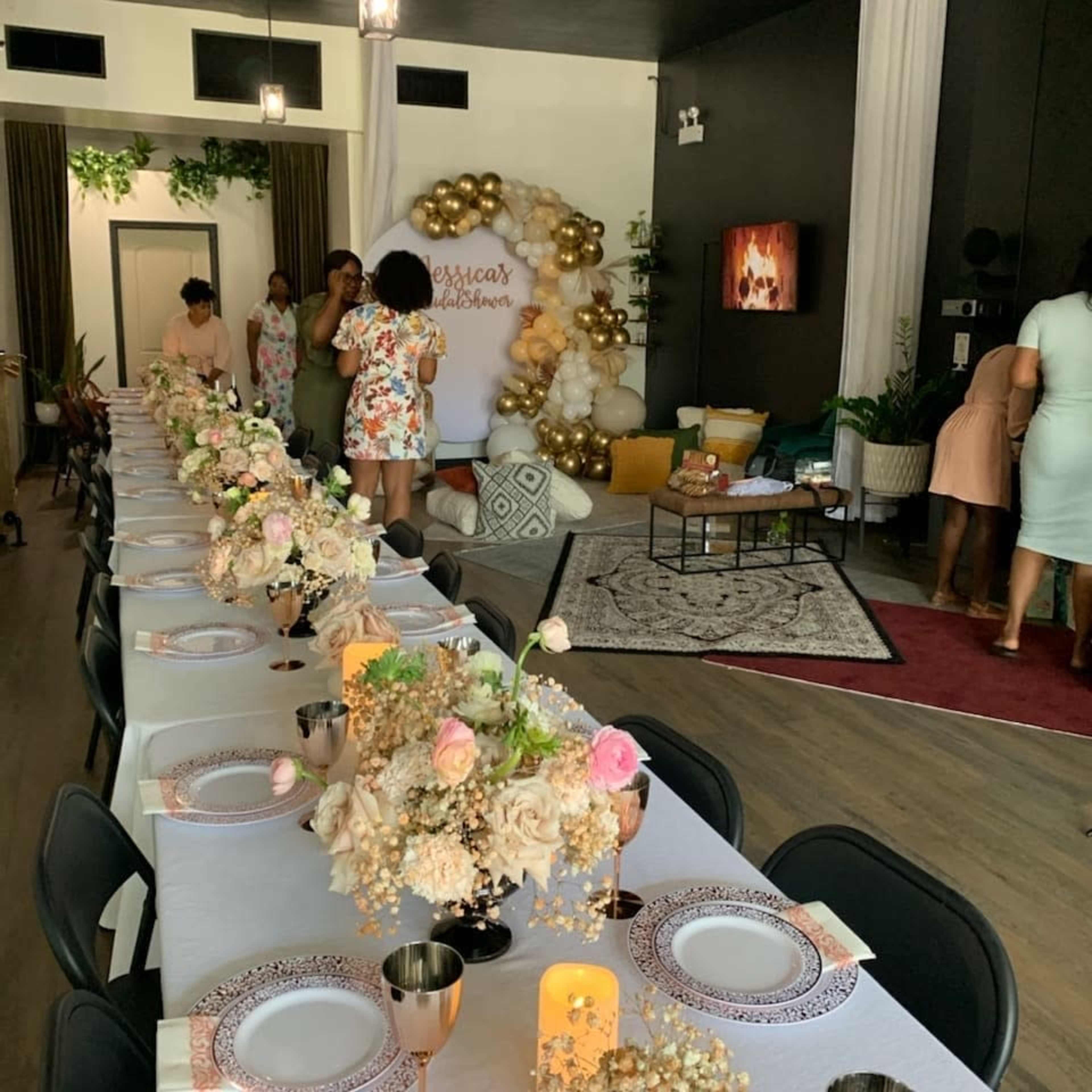 A decorated indoor event space with a long dining table set with floral centerpieces and guests preparing in the background.