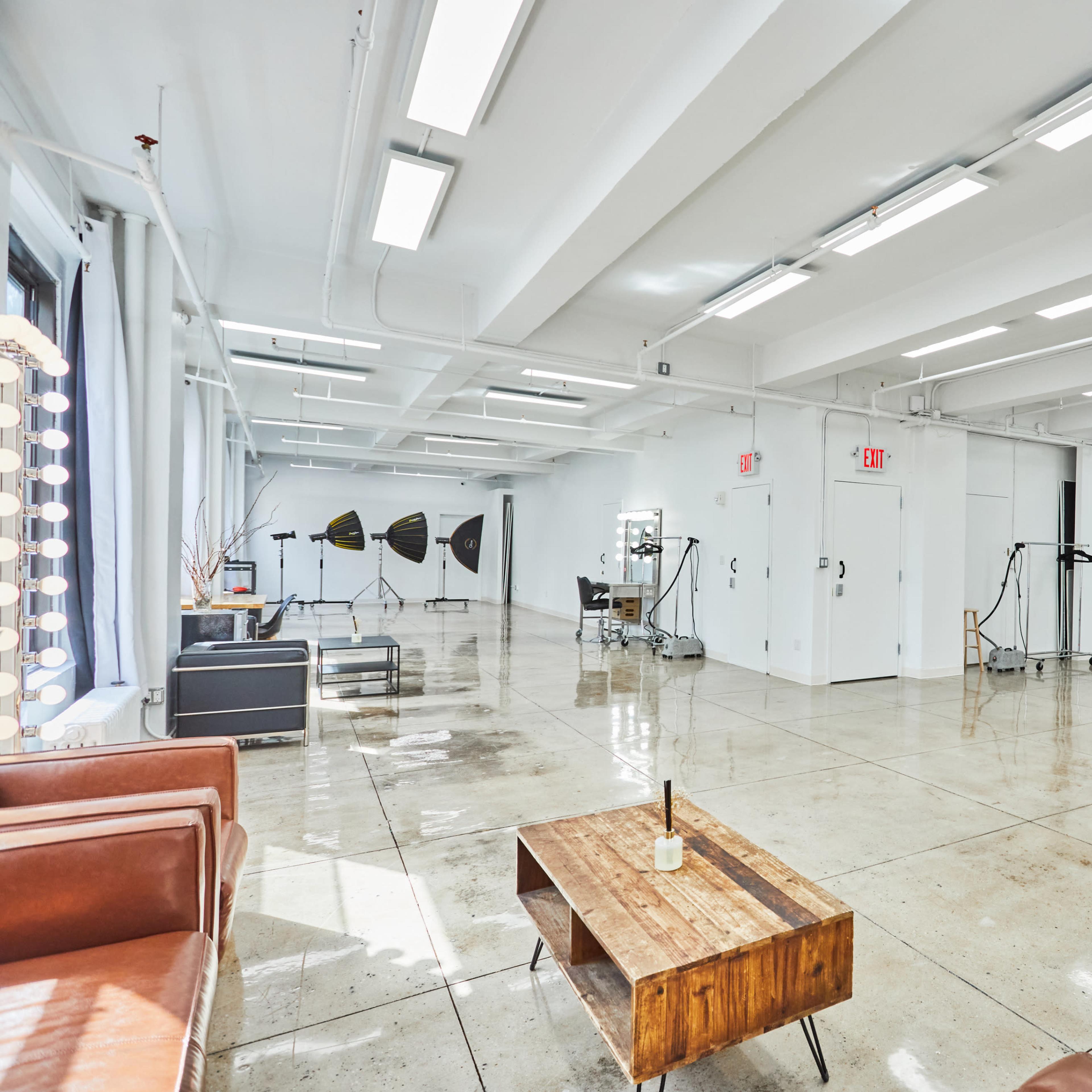 A spacious, well-lit studio with polished concrete floors, featuring several photography lighting setups and a modern lounge area with a wooden coffee table.