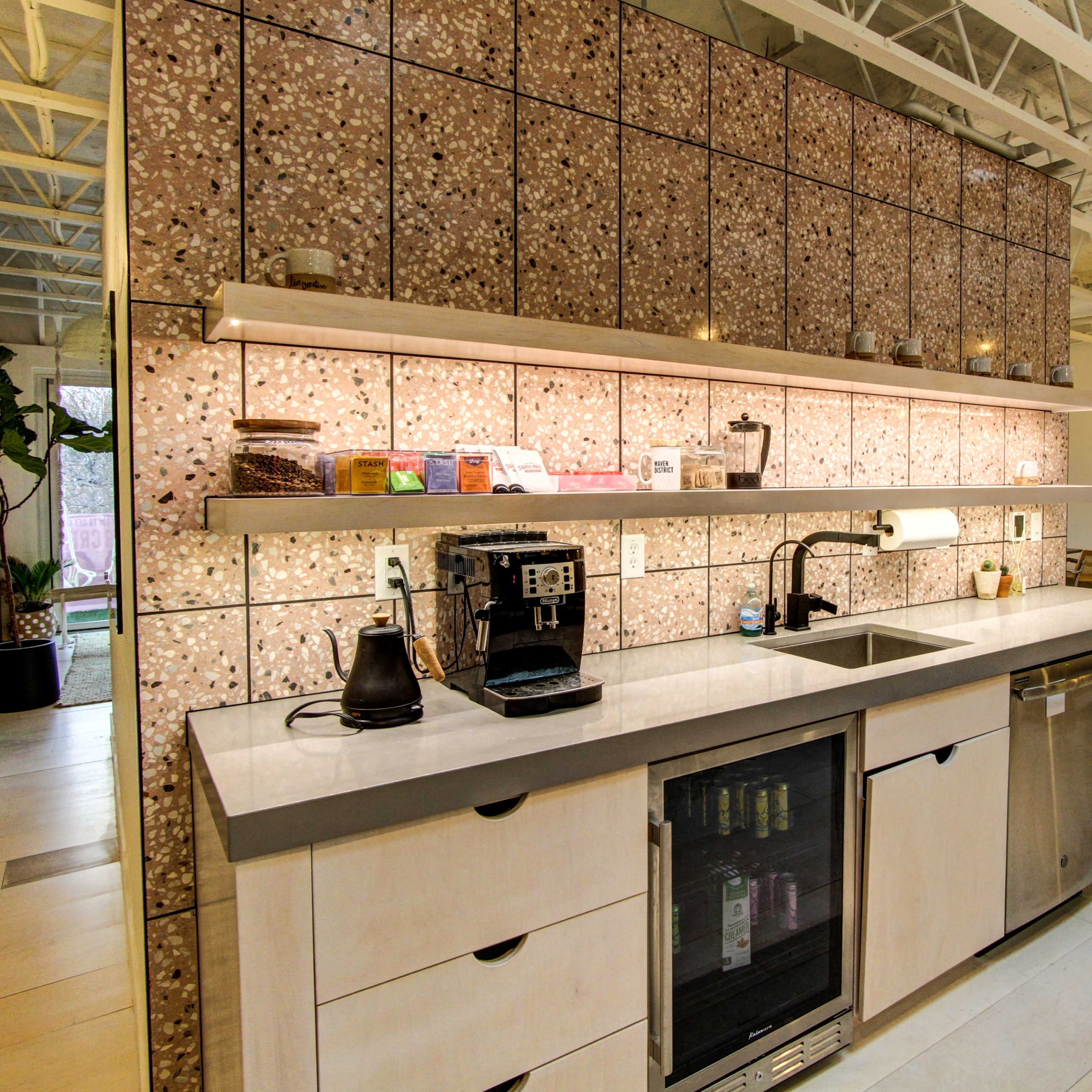 A modern kitchen with light wood cabinetry, a sink, a coffee machine, and a refrigerator, all set against a wall covered with textured stone tiles and ample overhead storage.