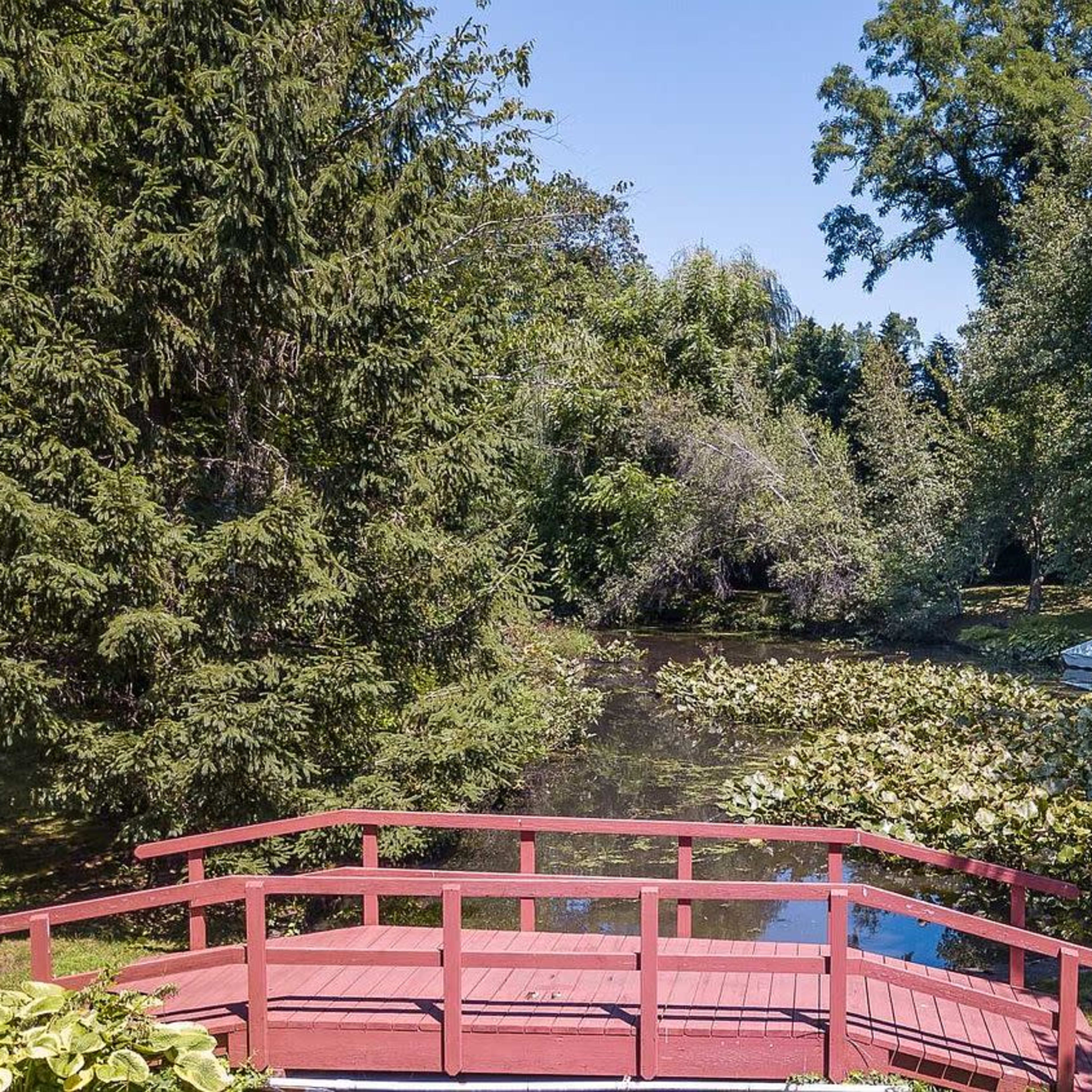 A red wooden bridge spans a small pond surrounded by lush greenery and a white boat rests at the water's edge.