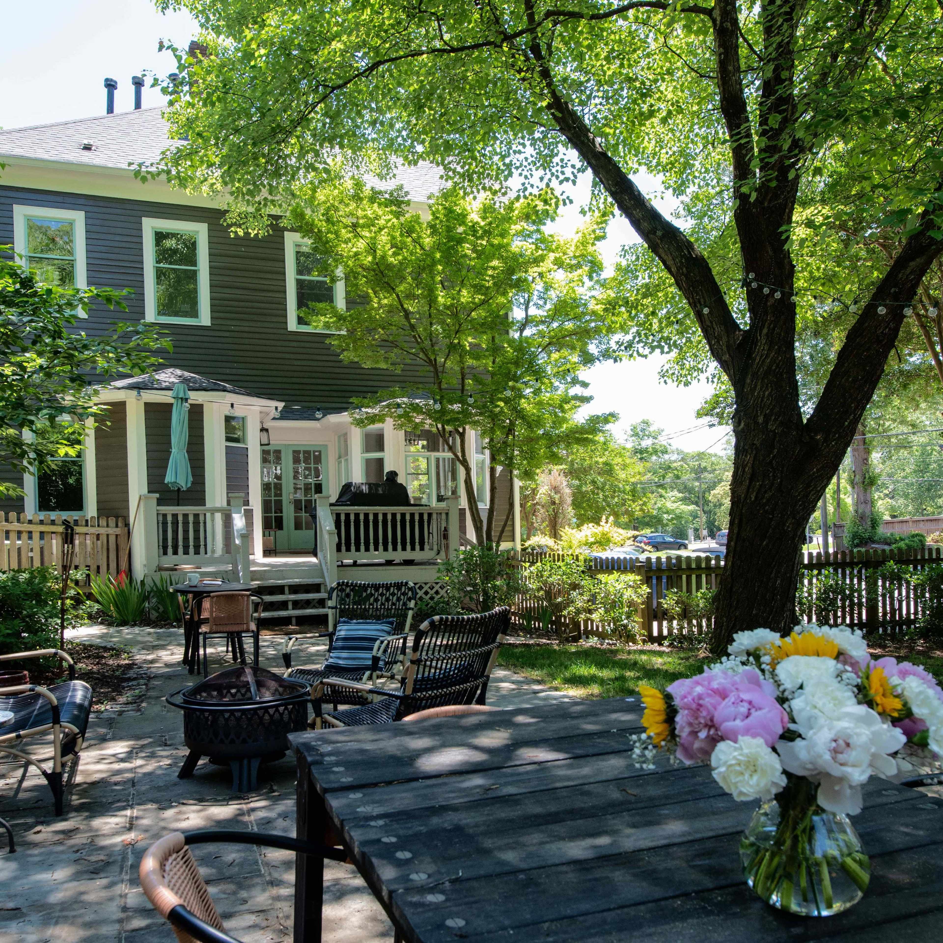 A spacious backyard features a wooden table with a vase of flowers, surrounded by chairs and overlooking a two-story house with large trees and a deck.