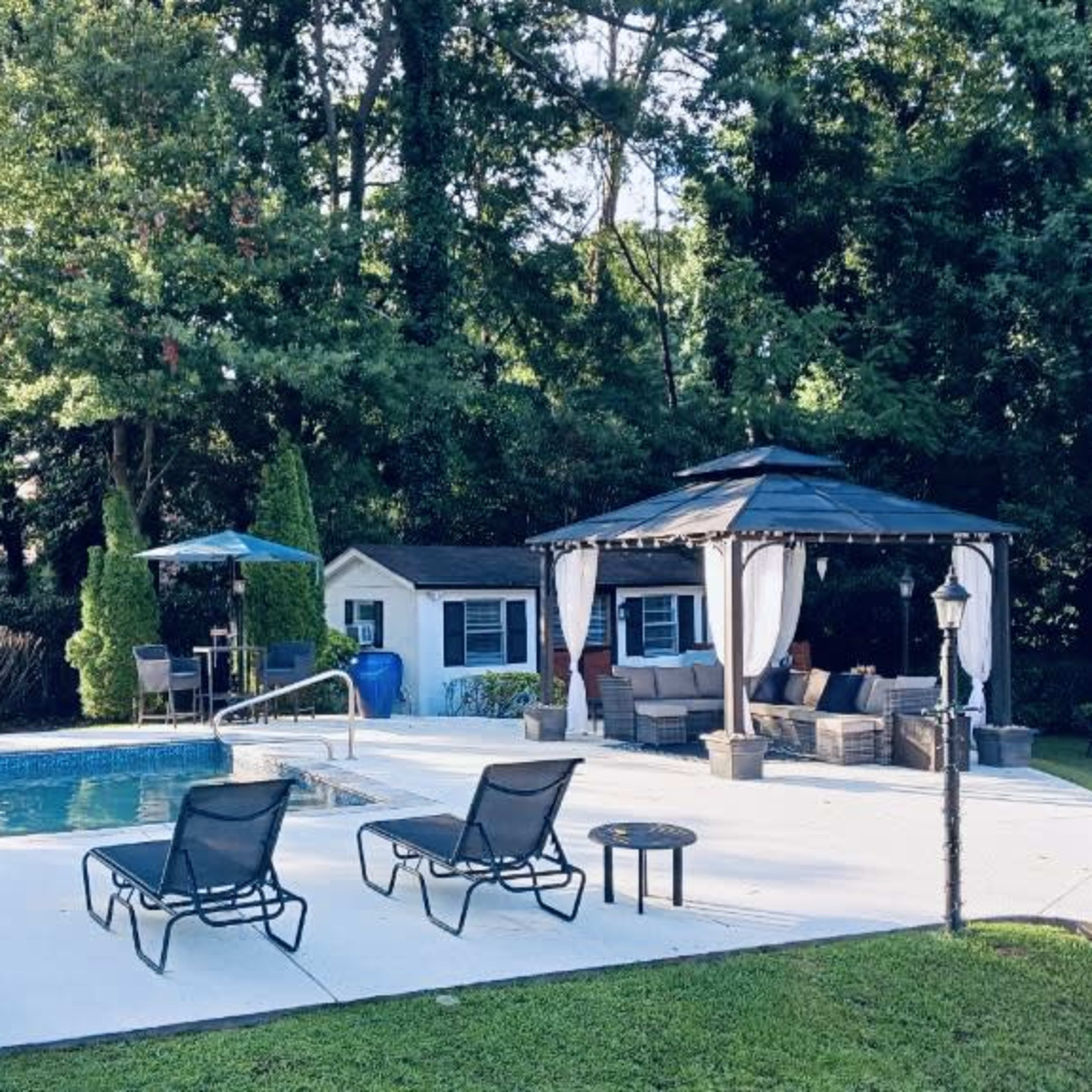A backyard with a swimming pool, a gazebo, lounge chairs, and a well-maintained lawn surrounded by trees.