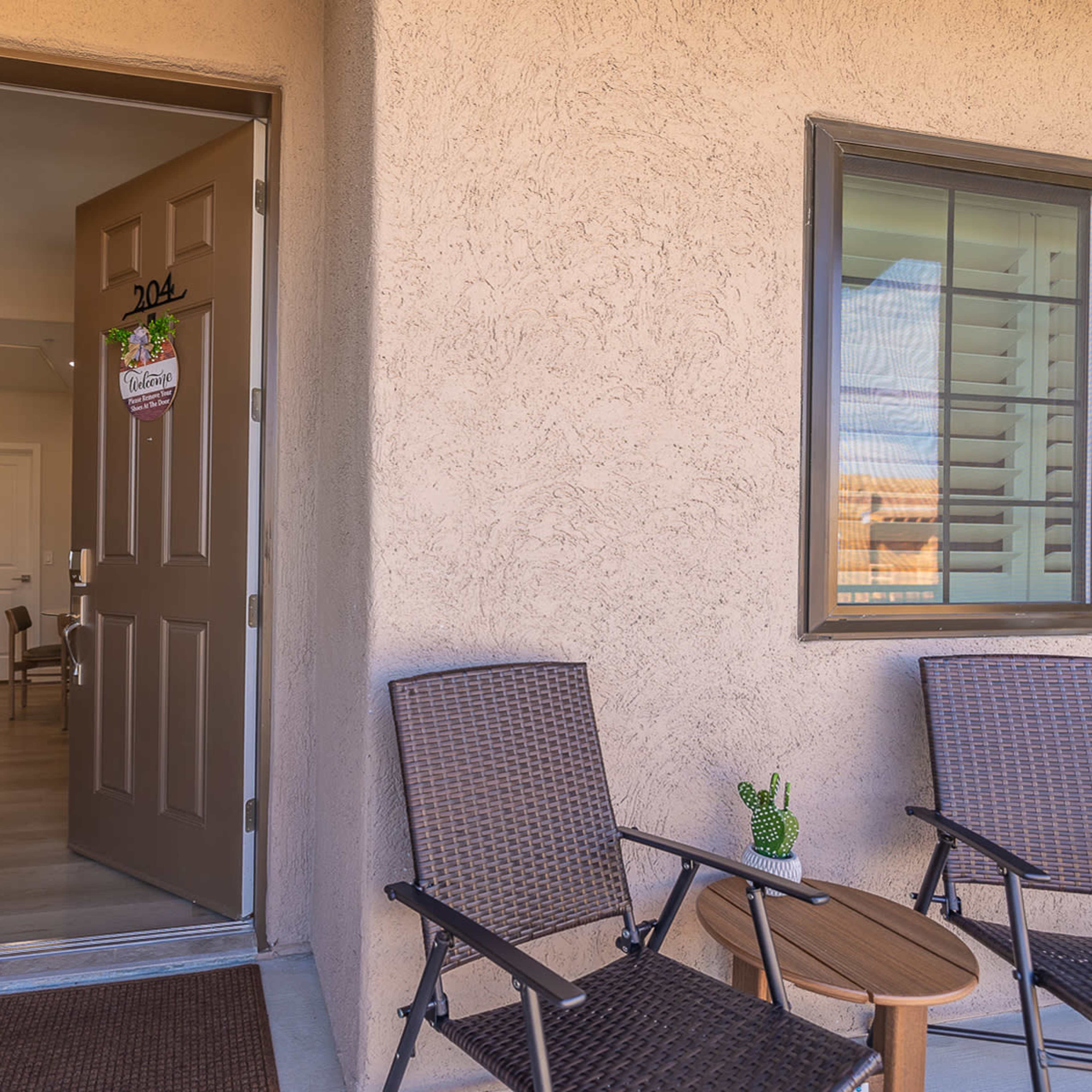 The entrance of an apartment with an open door, revealing a hallway, and two chairs with a small table on the porch.