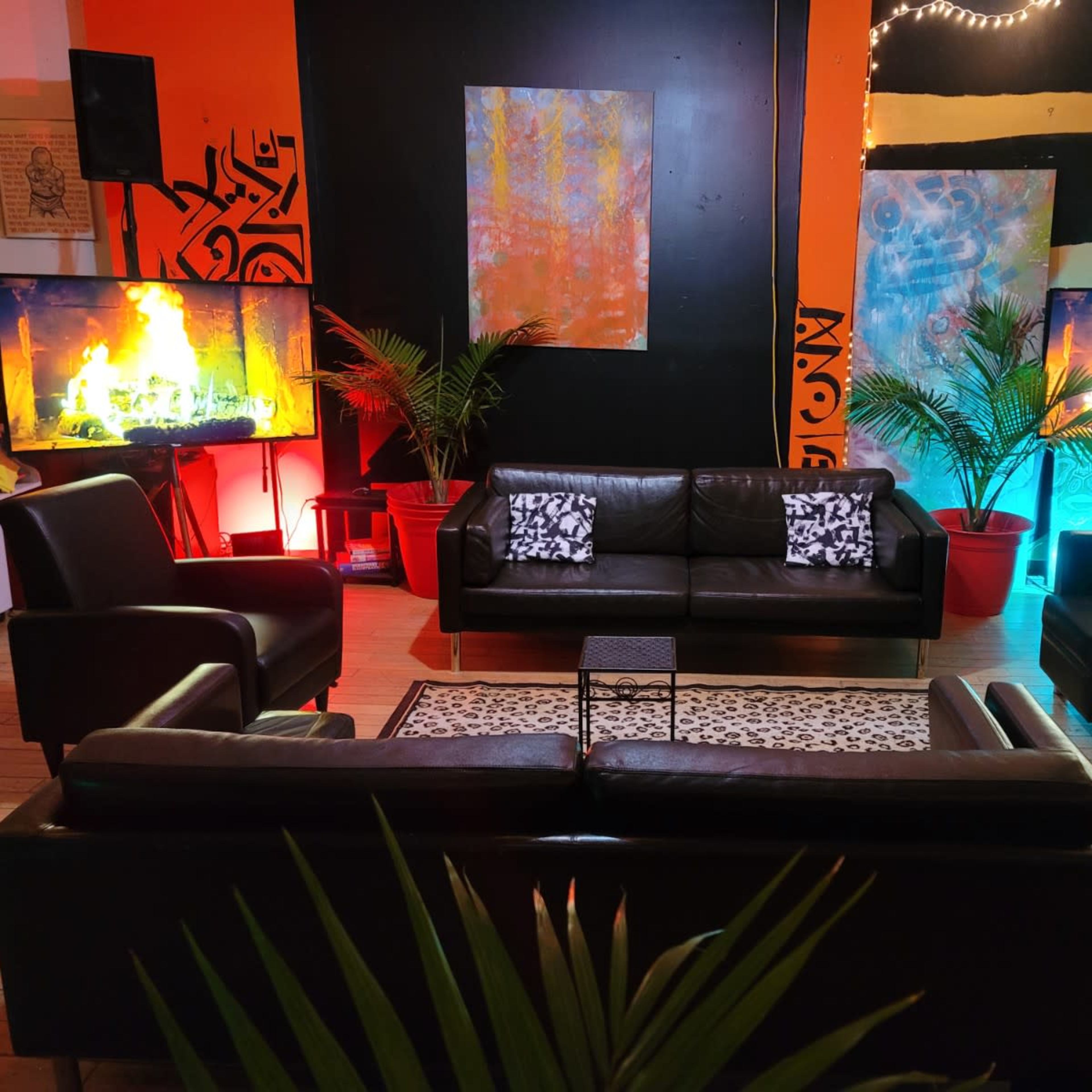 The scene features a modern lounge area with black leather furniture, two large televisions displaying flames, and colorful backgrounds illuminated by lights, complemented by potted plants.