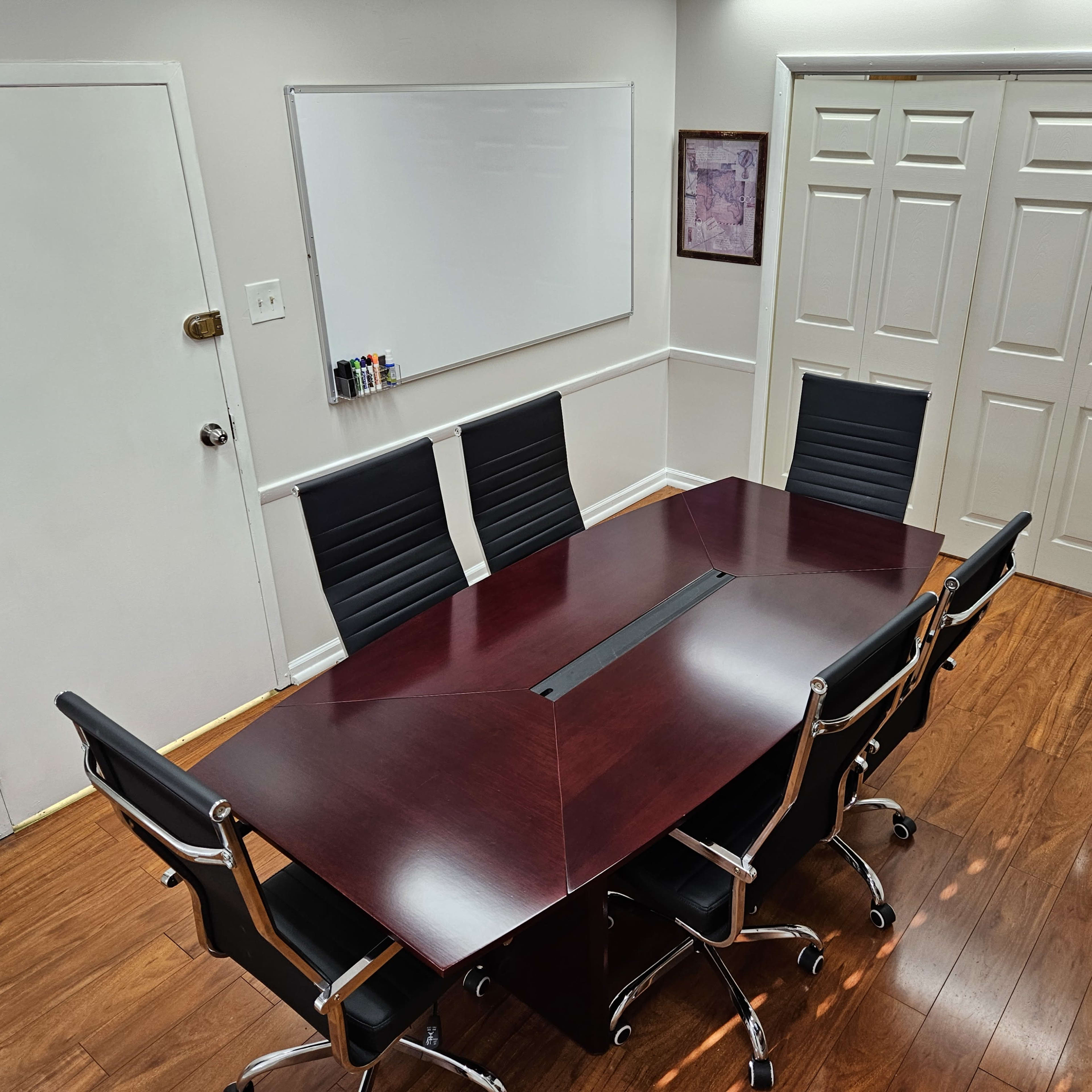 A conference room features a large, dark wooden table surrounded by six black office chairs, with a whiteboard and double doors in the background.