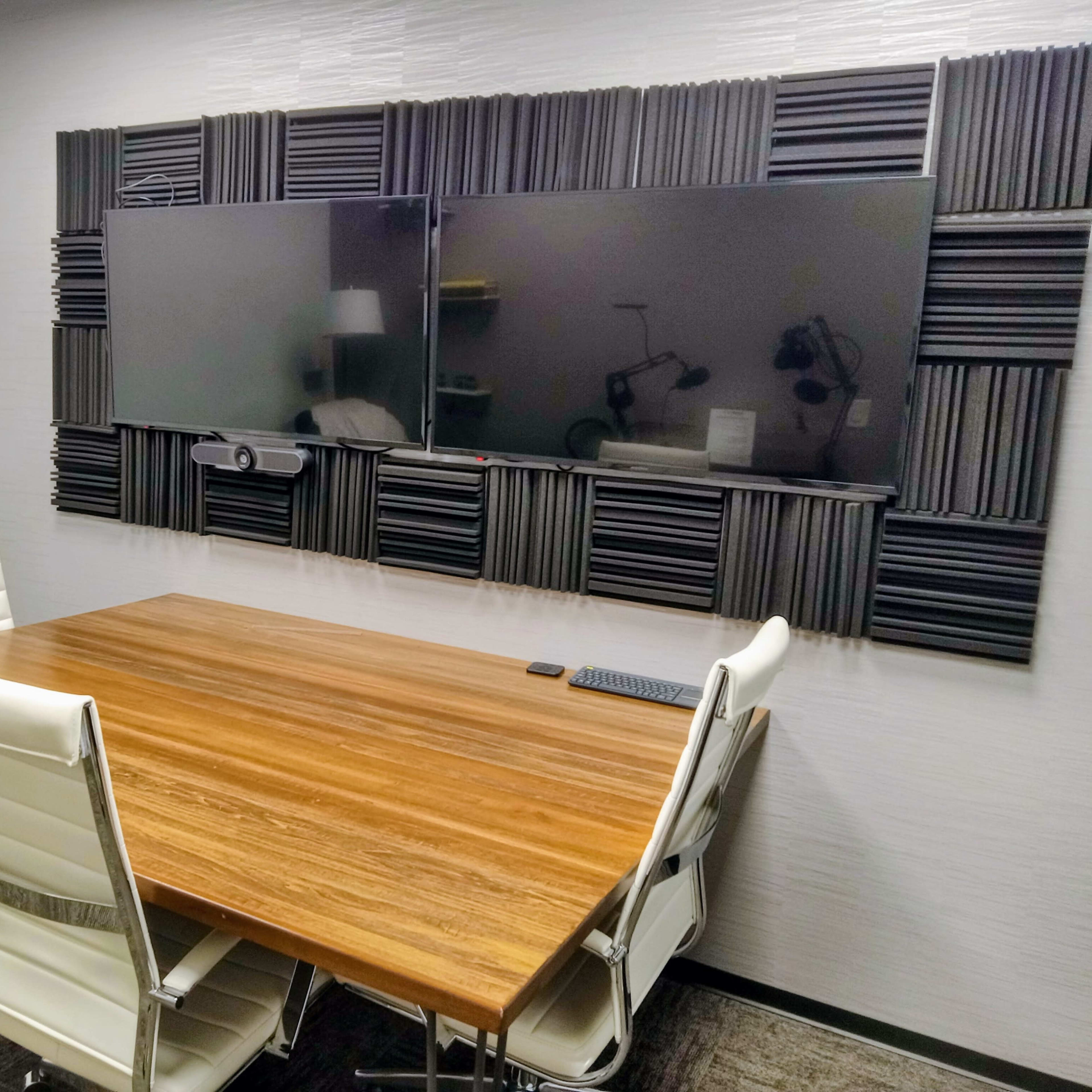 A modern conference room features a wooden table with white chairs and two large screens mounted on a wall with acoustic panels.