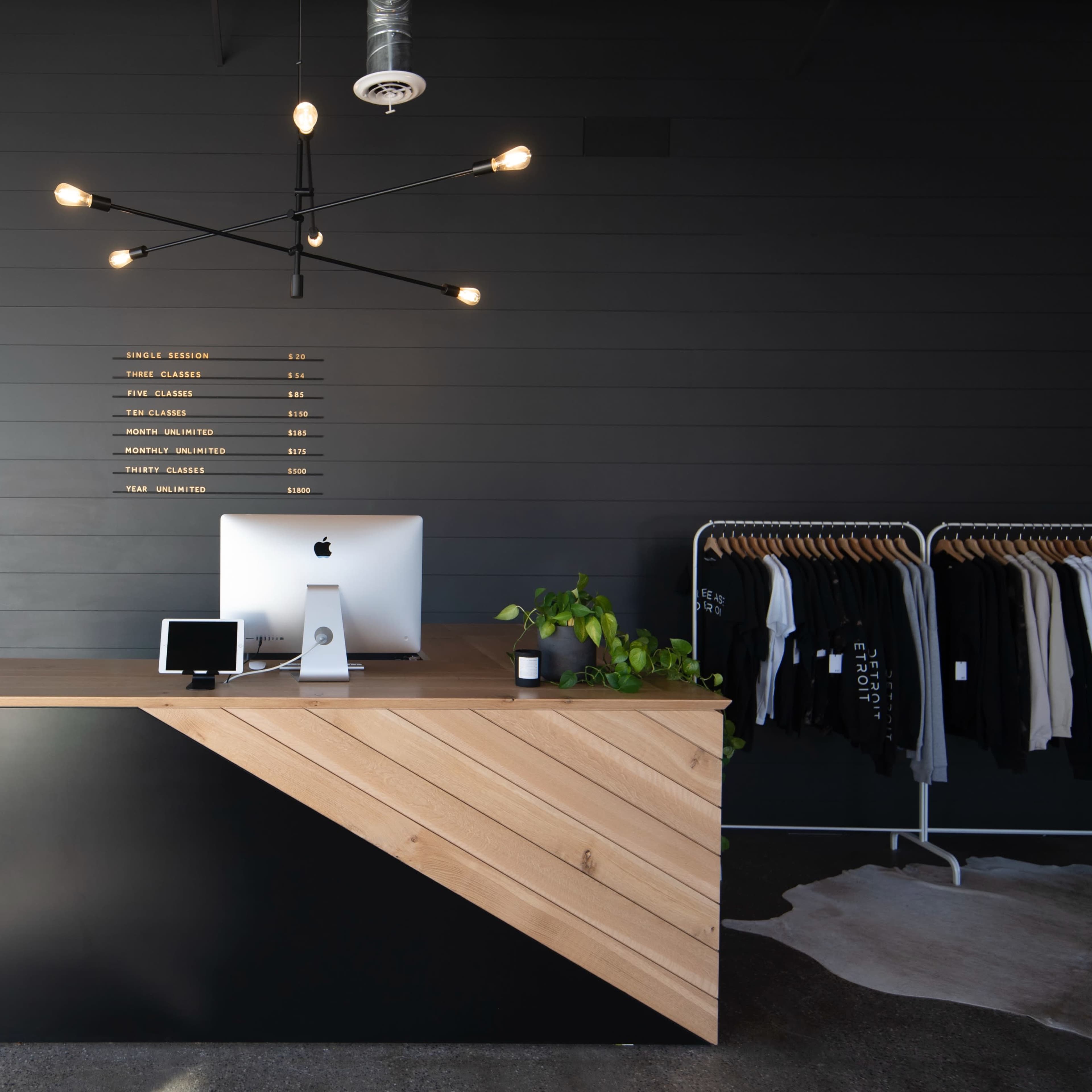 A modern reception area featuring a wooden reception desk with a computer, a plant, and clothing racks in a well-lit room with dark walls and decorative light fixtures.
