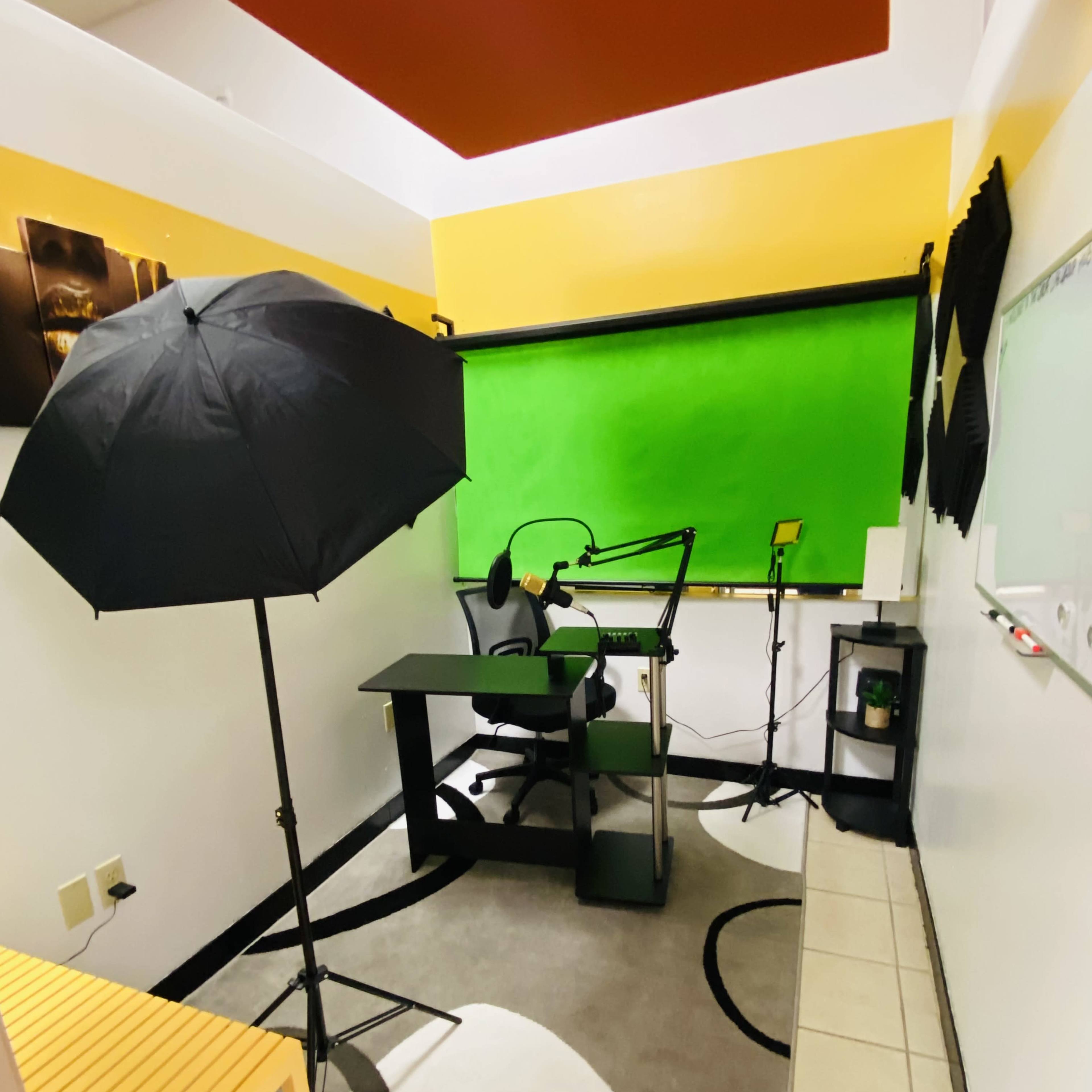 A recording studio with a black umbrella light, a microphone setup, a desk with a chair, and a green backdrop.