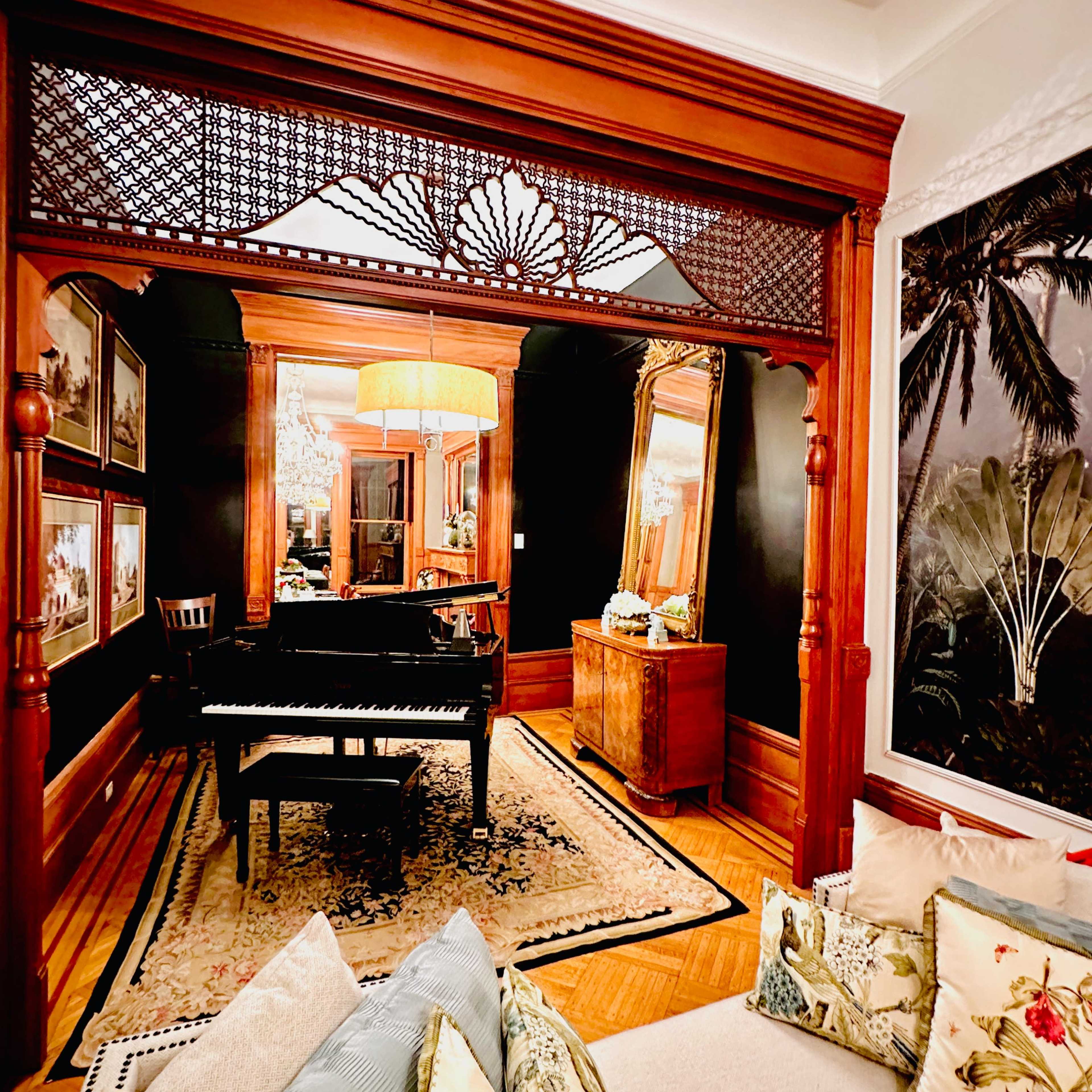 A cozy living space featuring a grand piano, decorative wooden architecture, and a large mirror, with a sofa adorned with floral cushions in the foreground.