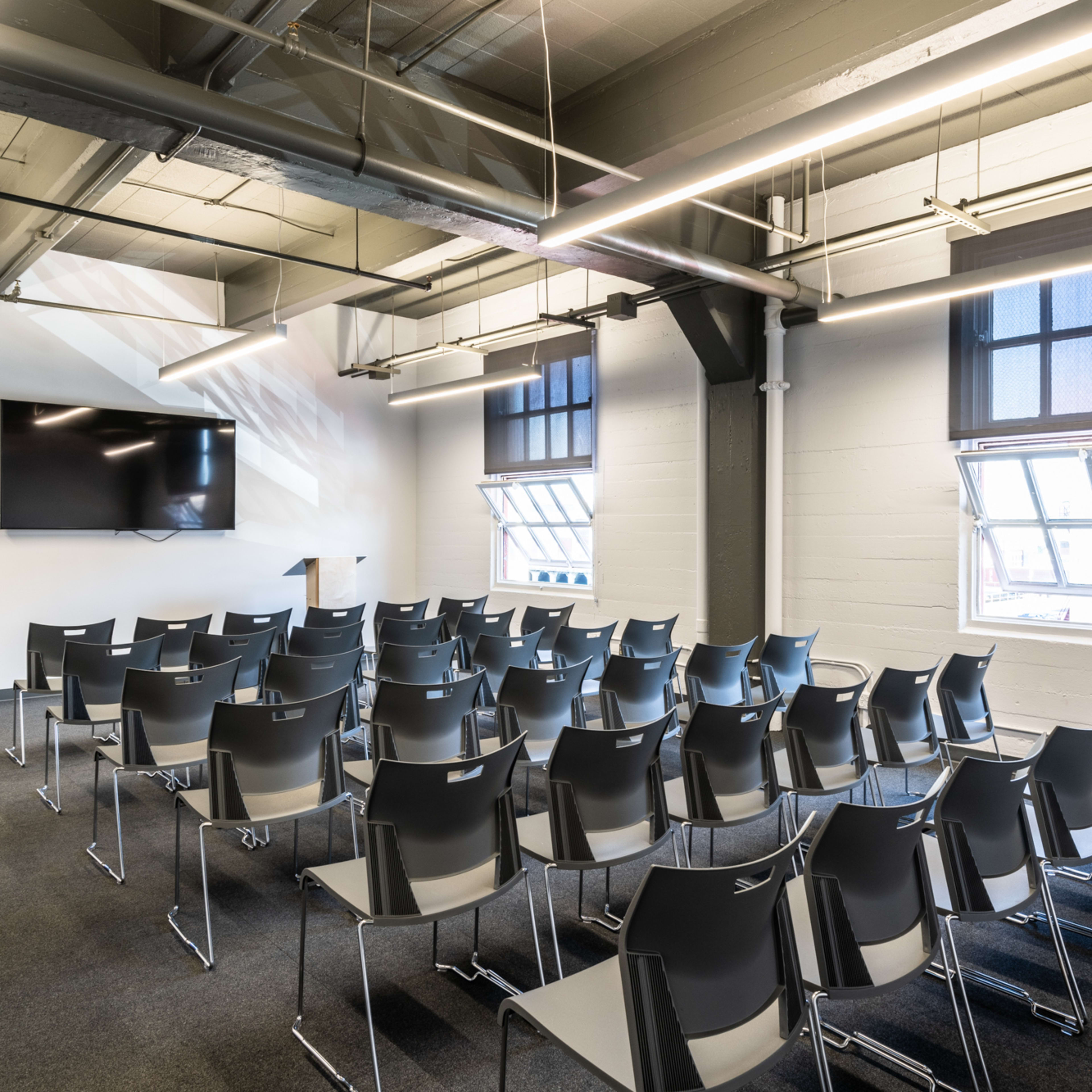 A conference room filled with rows of empty gray chairs, a large screen mounted on the wall, and windows that provide natural light.