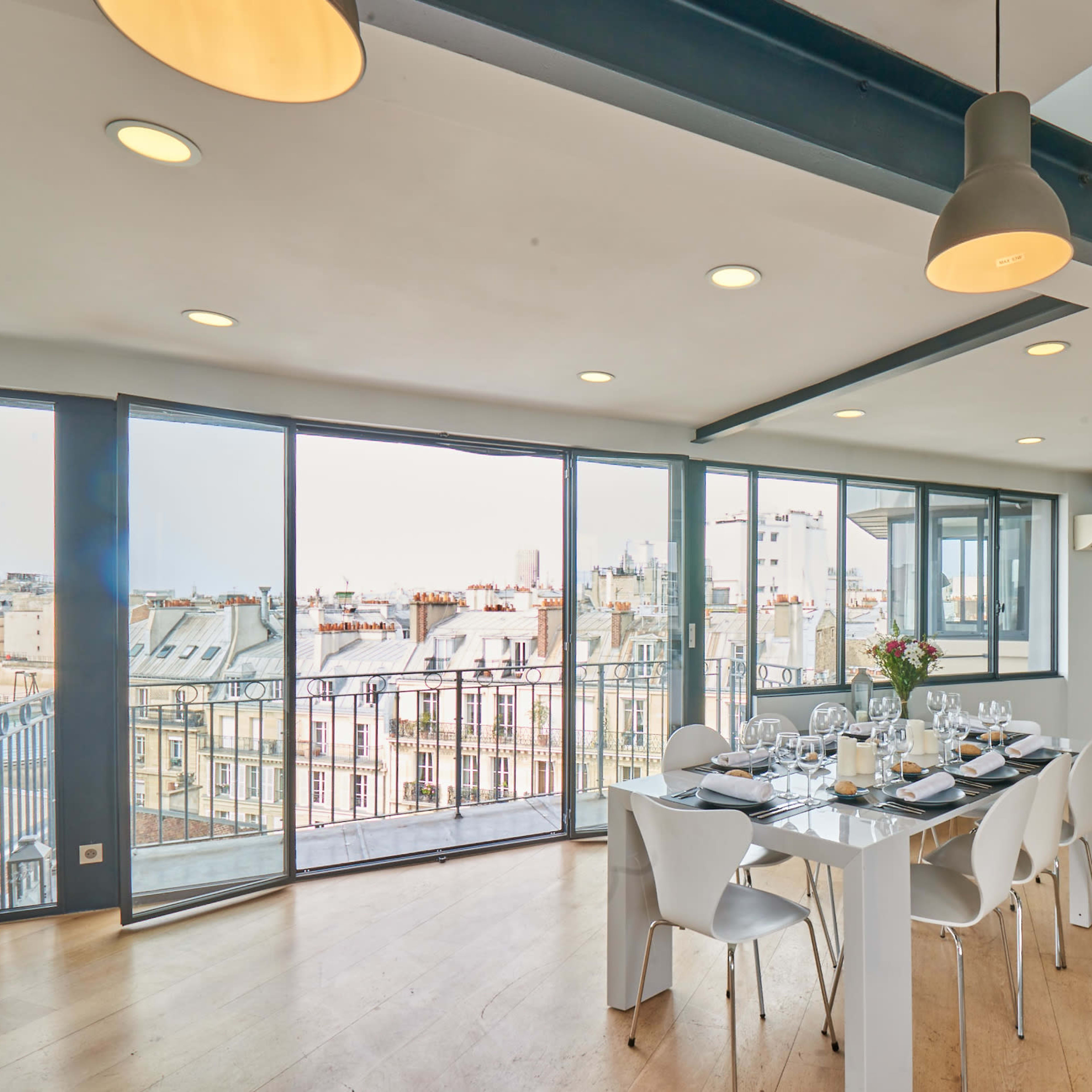 A modern dining area with a large table set for a meal, surrounded by large windows offering a view of the rooftops and skyline outside.
