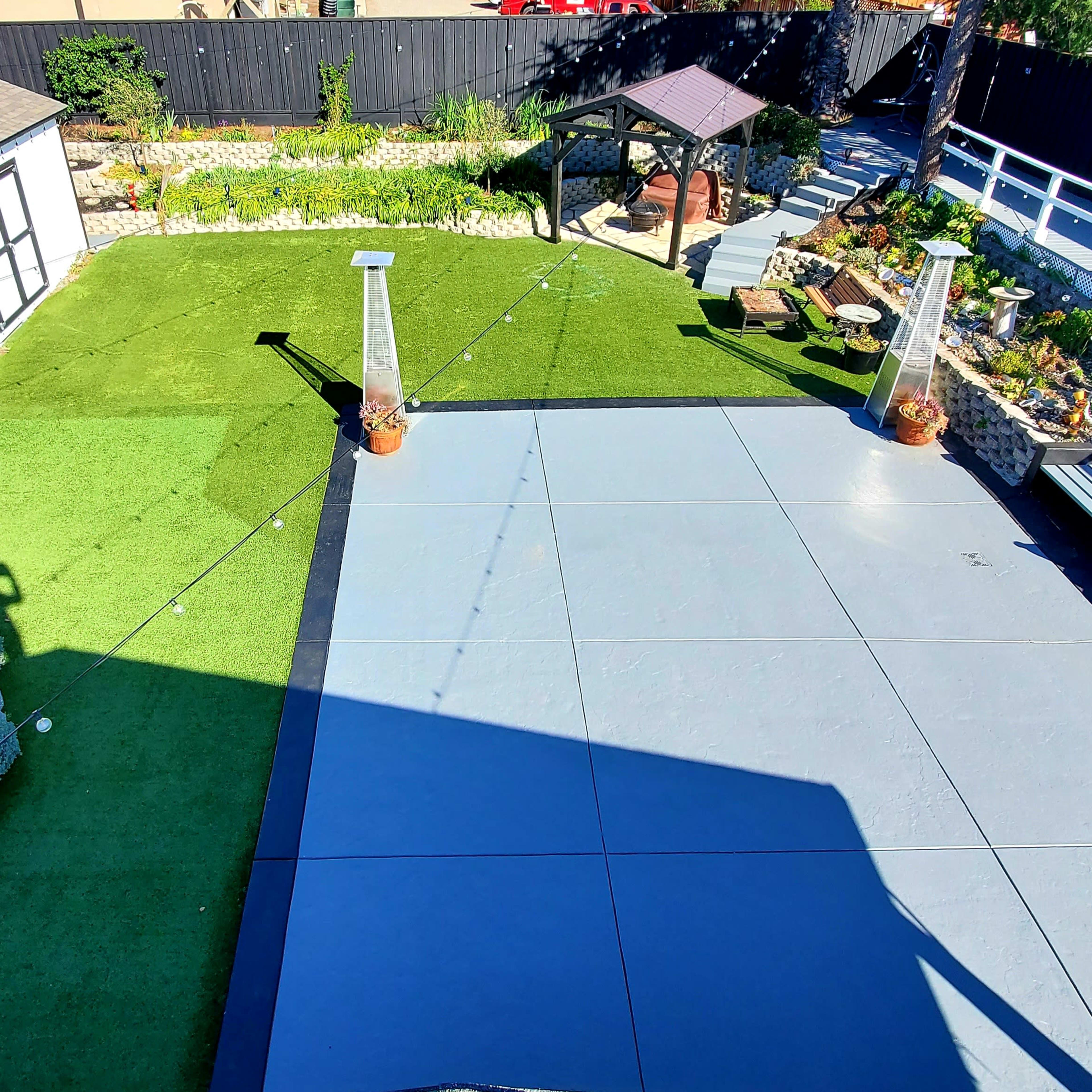 A landscaped outdoor area featuring a gray paving surface, artificial grass, a gazebo, and various garden features including potted plants and a small shed.