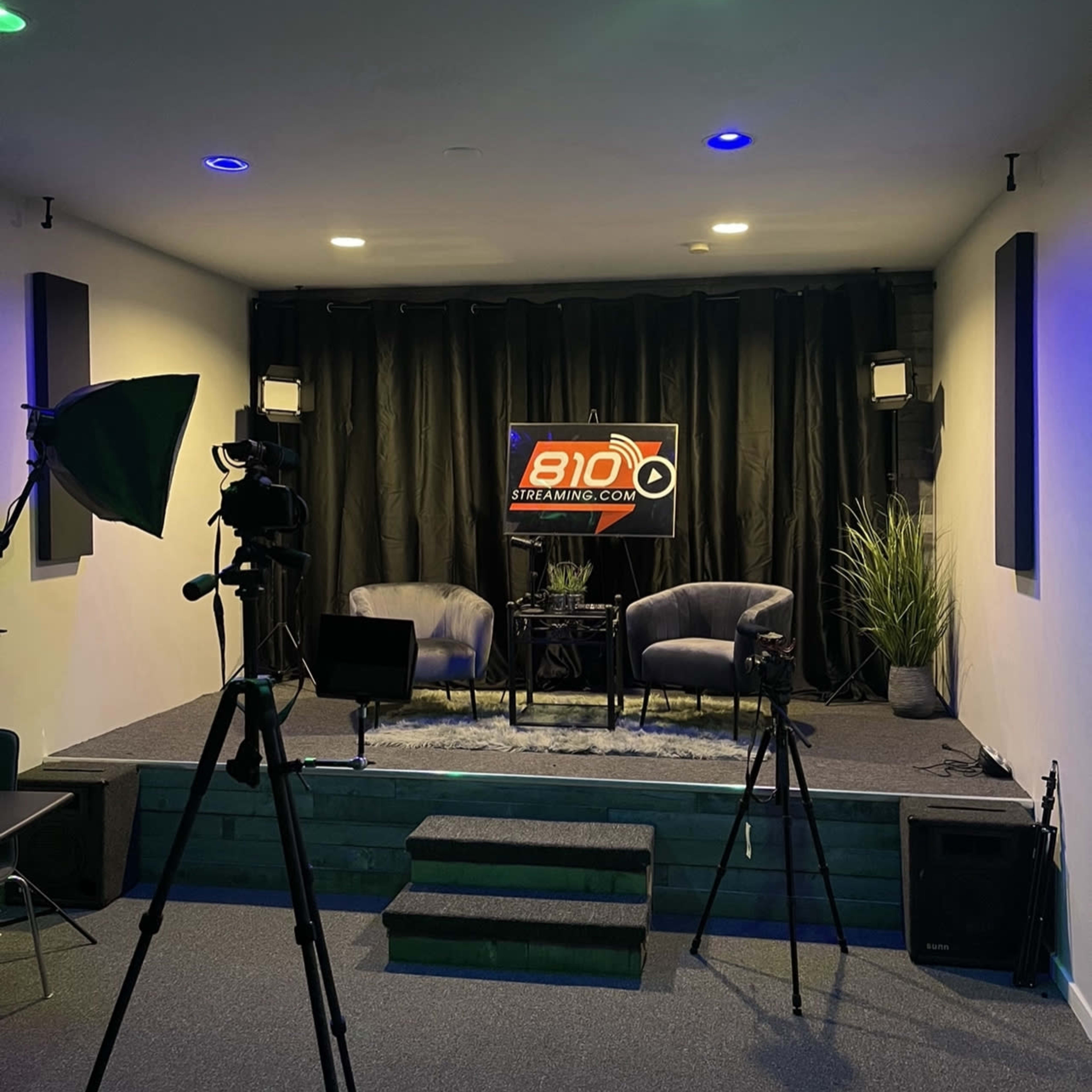 A streaming studio with two chairs, a small table, and lighting equipment, set up on a raised stage with a large backdrop displaying a logo.