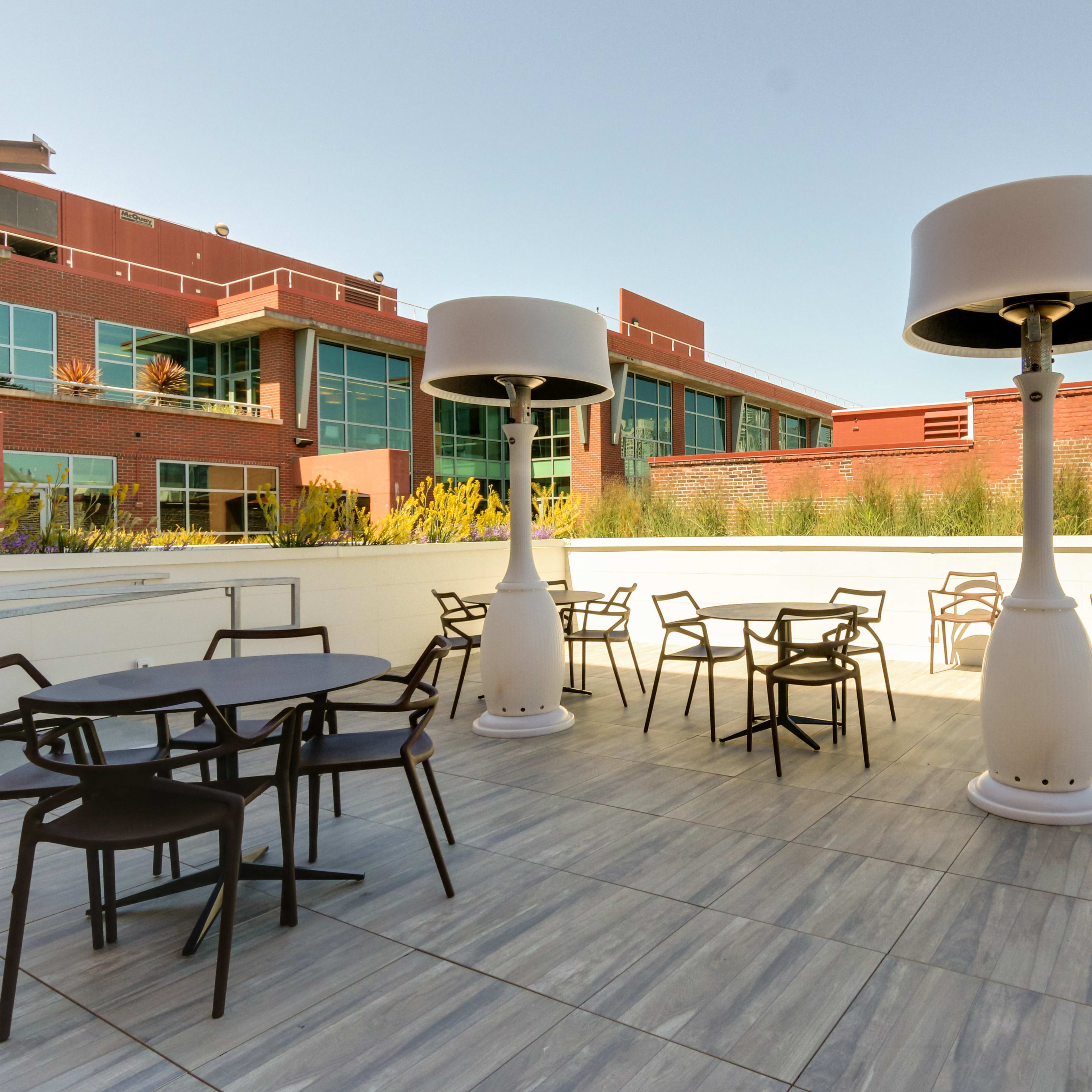 An outdoor patio area with several tables and chairs, two large heaters, and buildings in the background.
