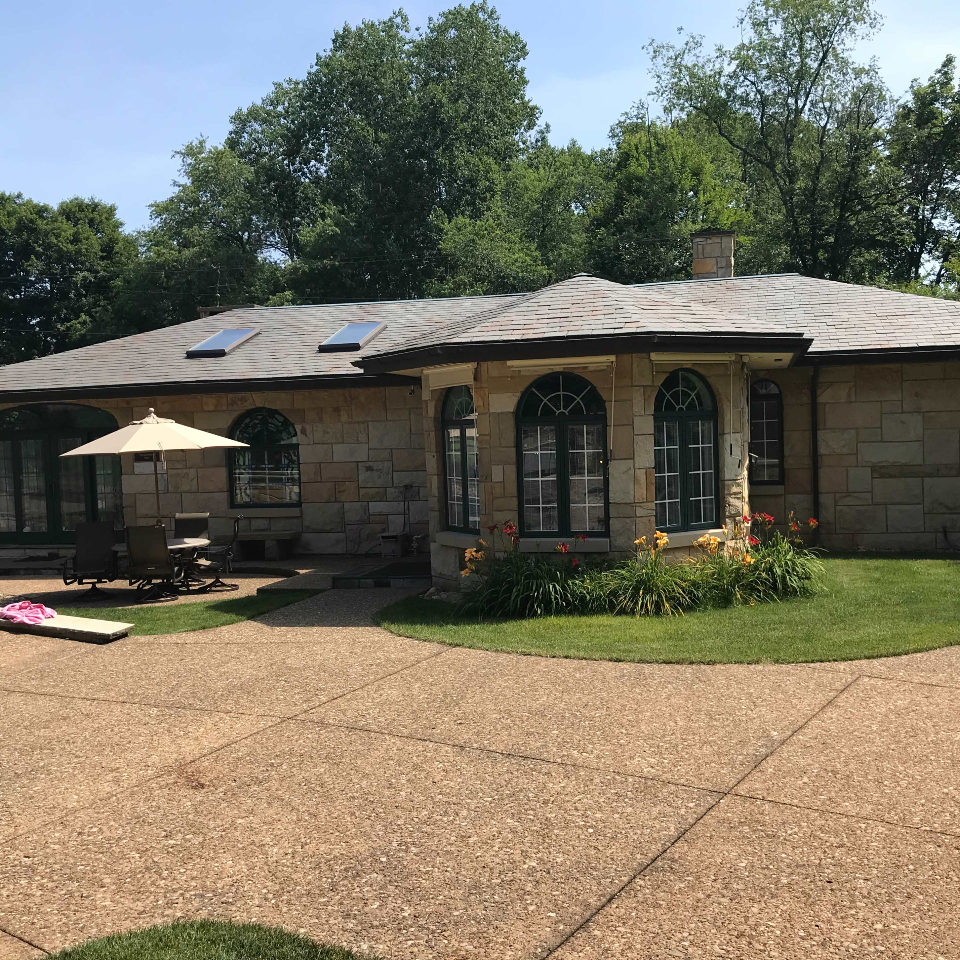 A stone house with large arched windows, surrounded by well-maintained grass and landscaping, with a patio umbrella and outdoor seating area.