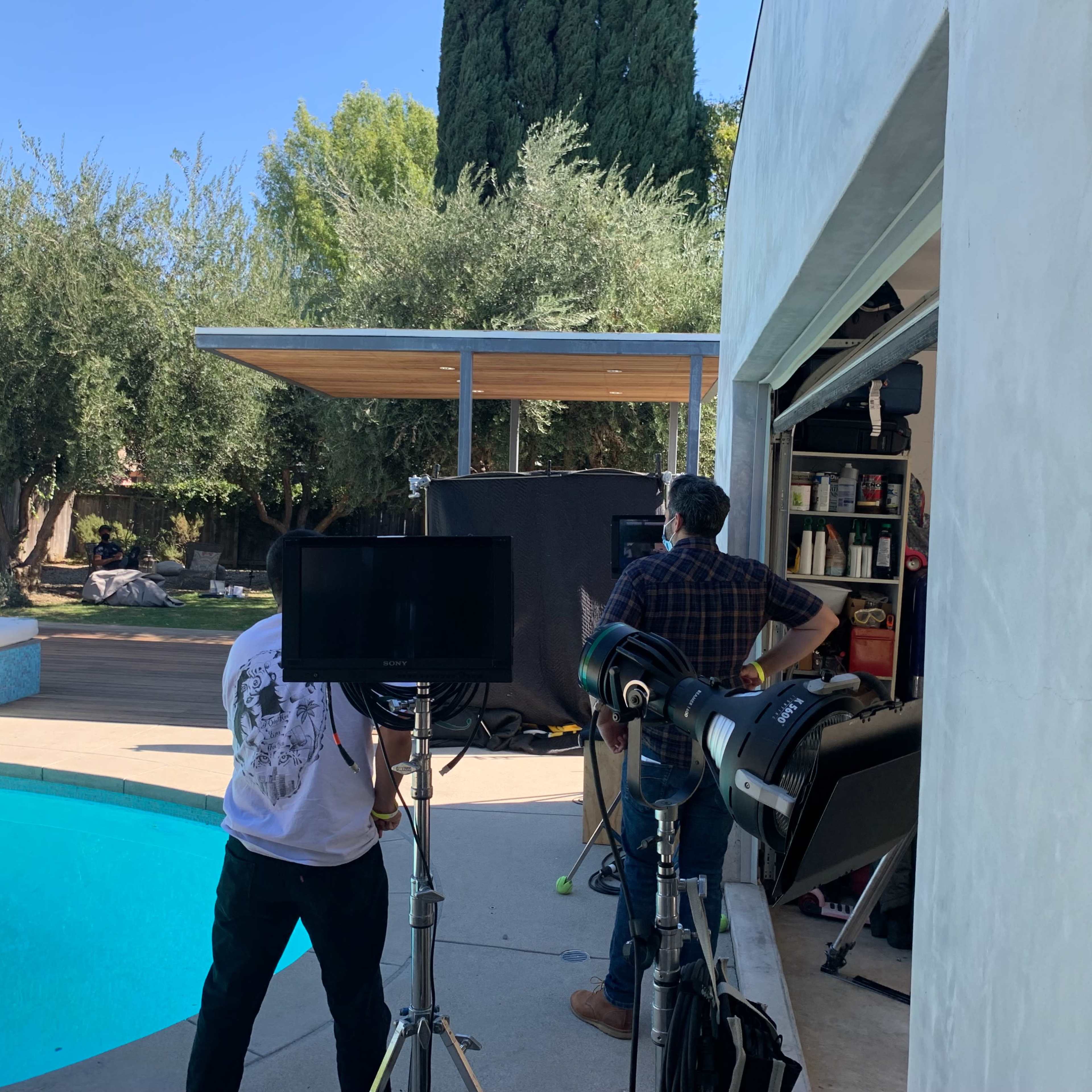 Two people set up filming equipment near a swimming pool in a backyard.