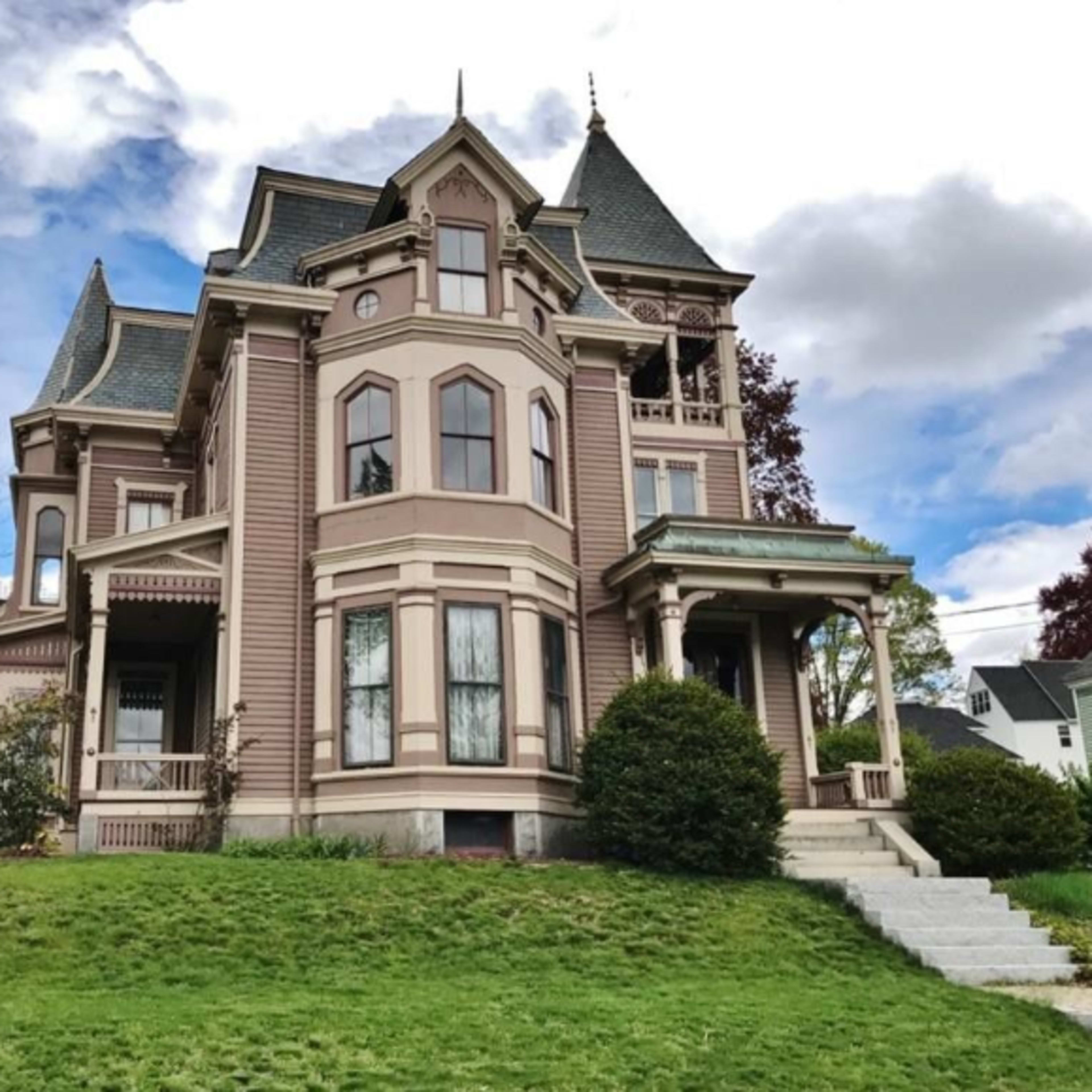 A large Victorian-style house with a steep roof and ornate details is situated on a green lawn.