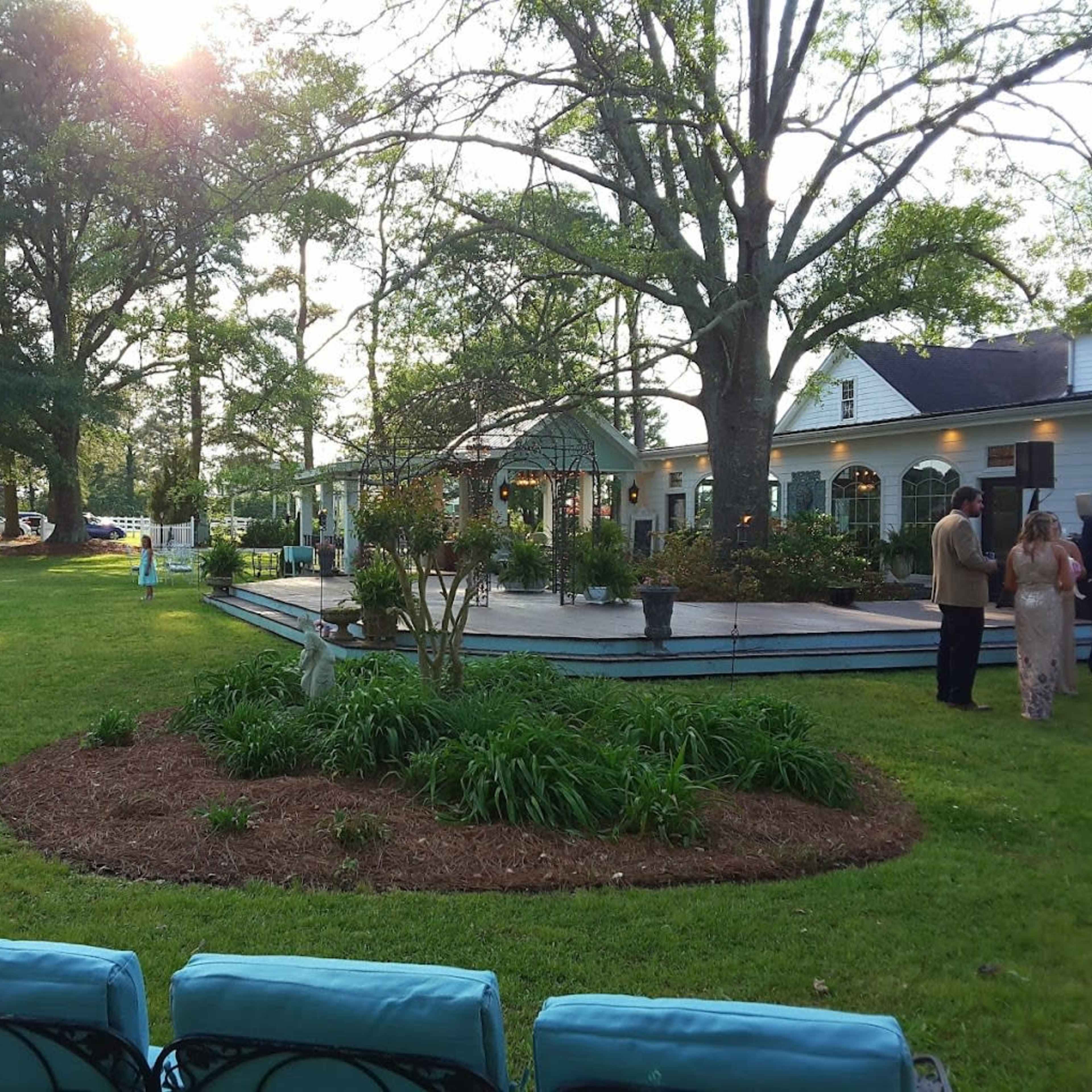 A landscaped area with plush green grass and a circular flower bed, featuring a spacious building and people gathered on a wooden deck in the background, framed by large trees and a soft sunset.