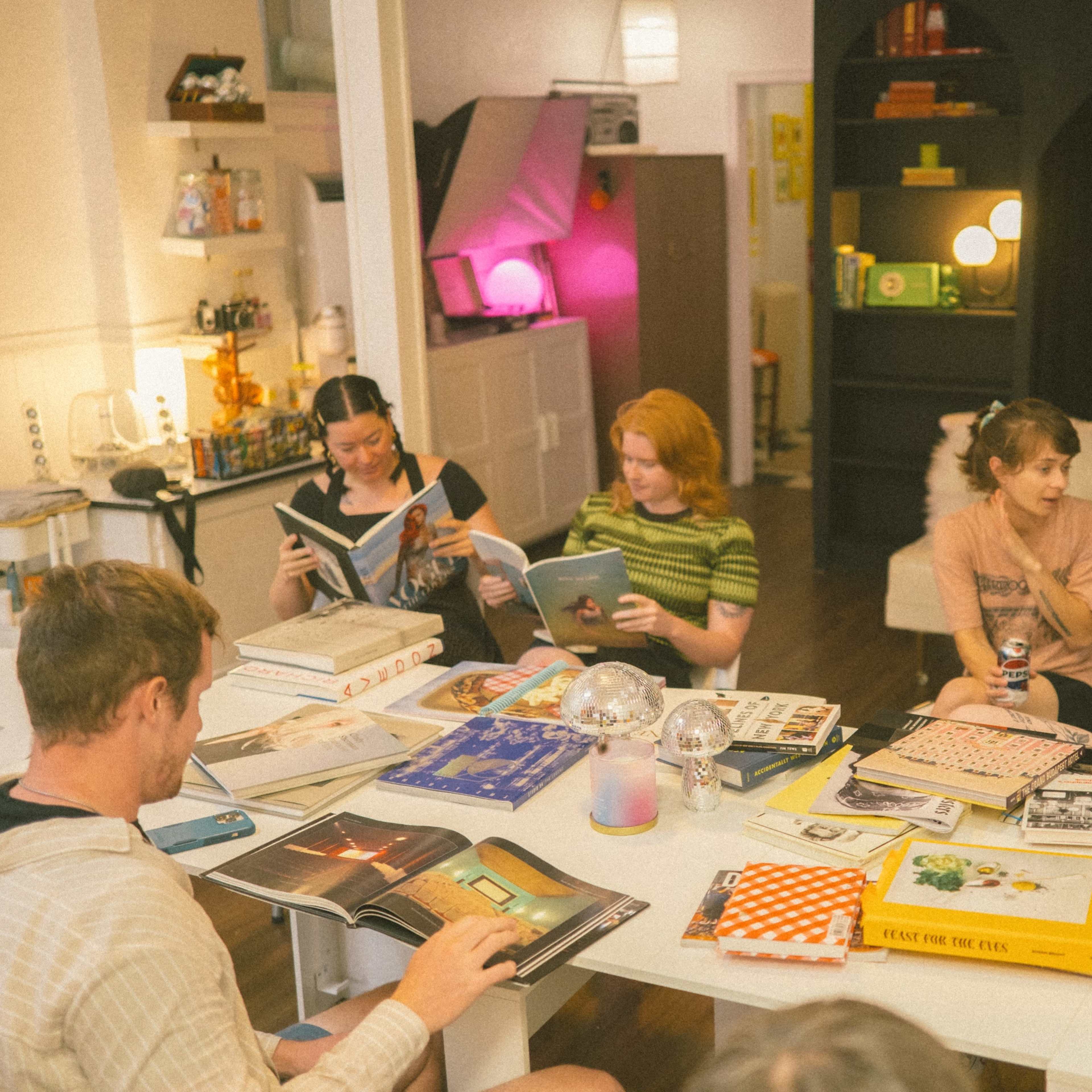 A group of people sits around a table in a well-lit room, engaging with various magazines and books.