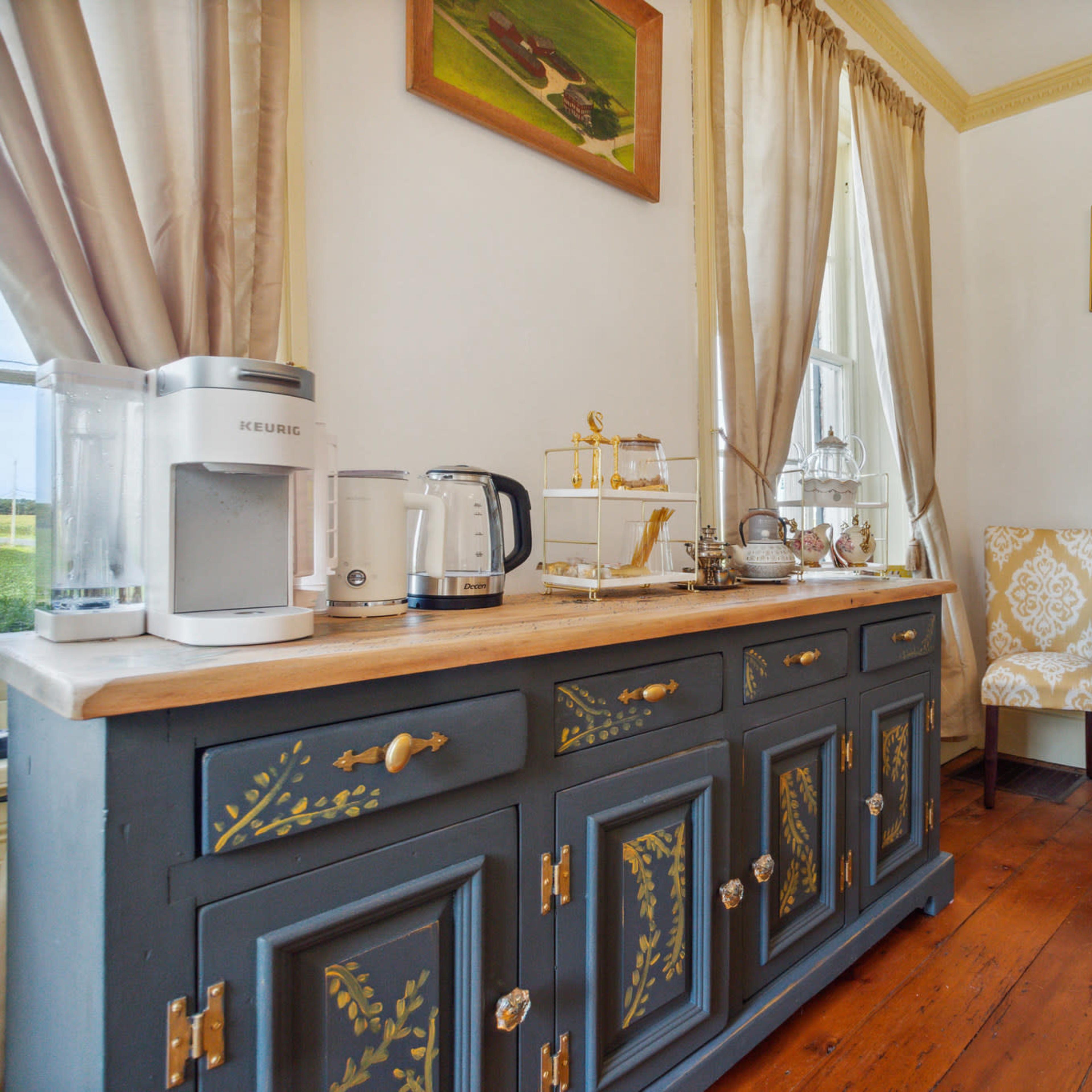 A cozy room features a wooden cabinet with dishes and a kettle, two patterned chairs, and artwork on the walls.