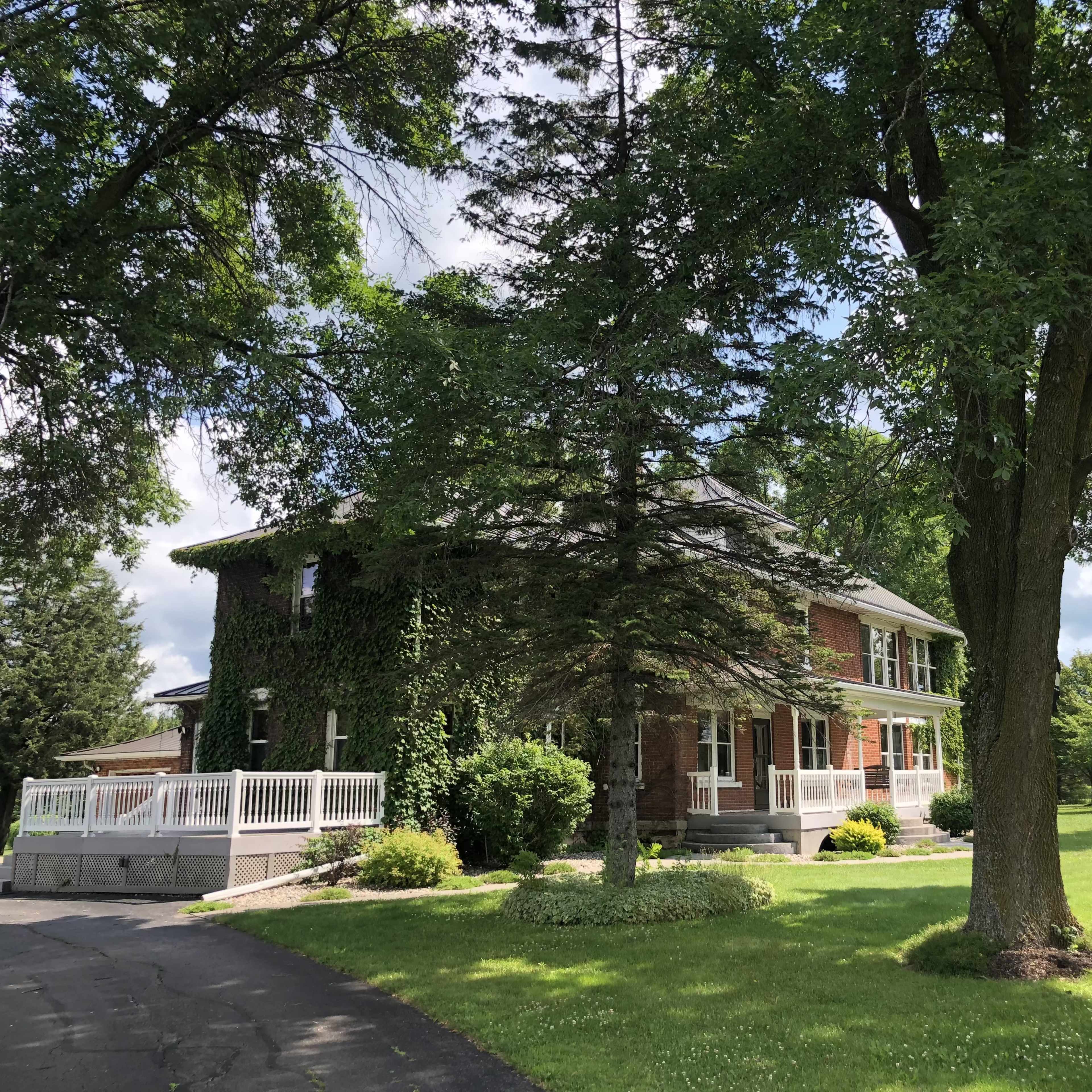 A brick house covered in ivy sits beside a large tree and manicured lawn under a partly cloudy sky.