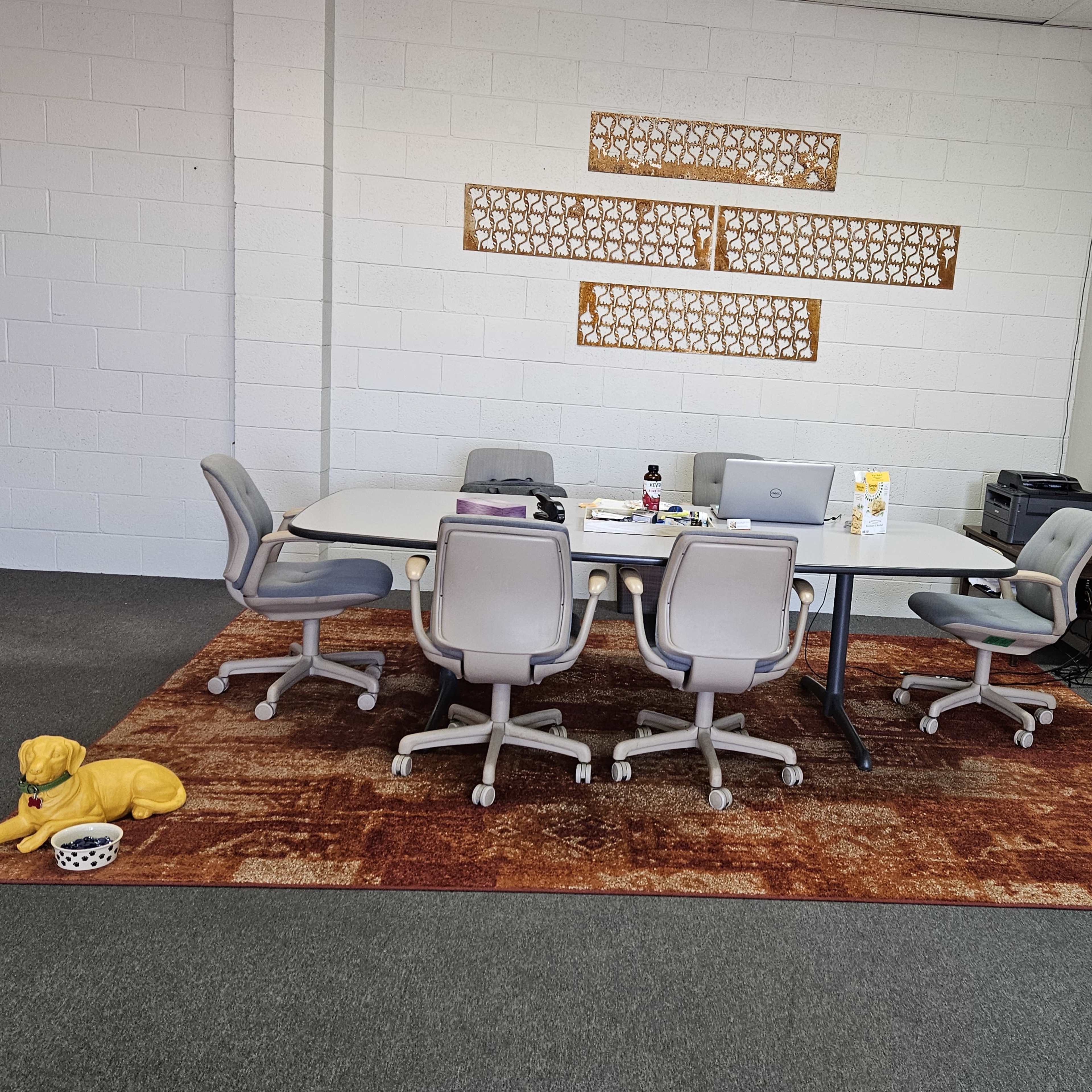 A meeting room features a large table surrounded by six gray chairs, a computer, and a decorative rug, with a dog lying on the floor beside a bowl.
