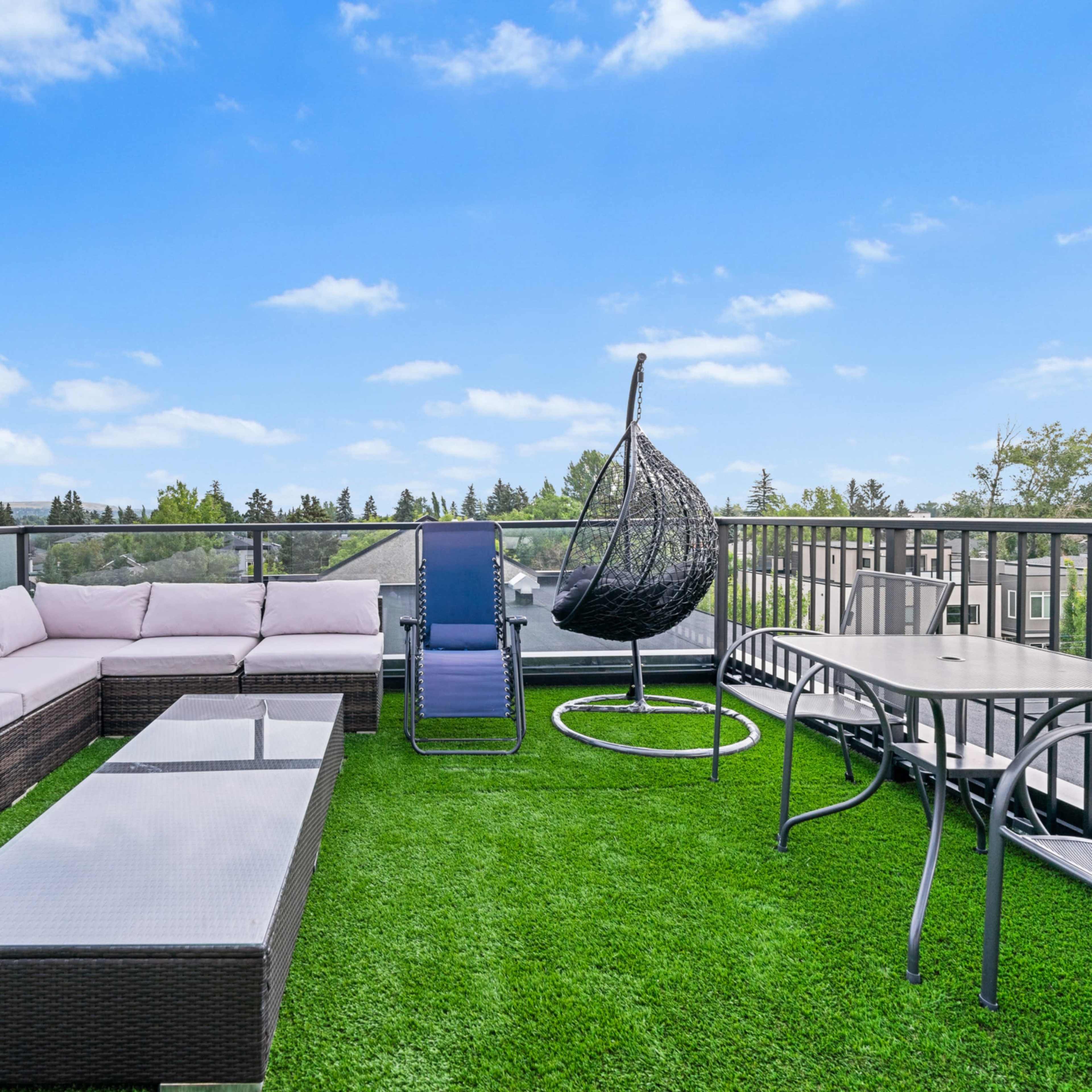A rooftop terrace features a wicker sectional sofa, a hanging chair, a lounge chair, and a dining table, all set on green artificial grass.