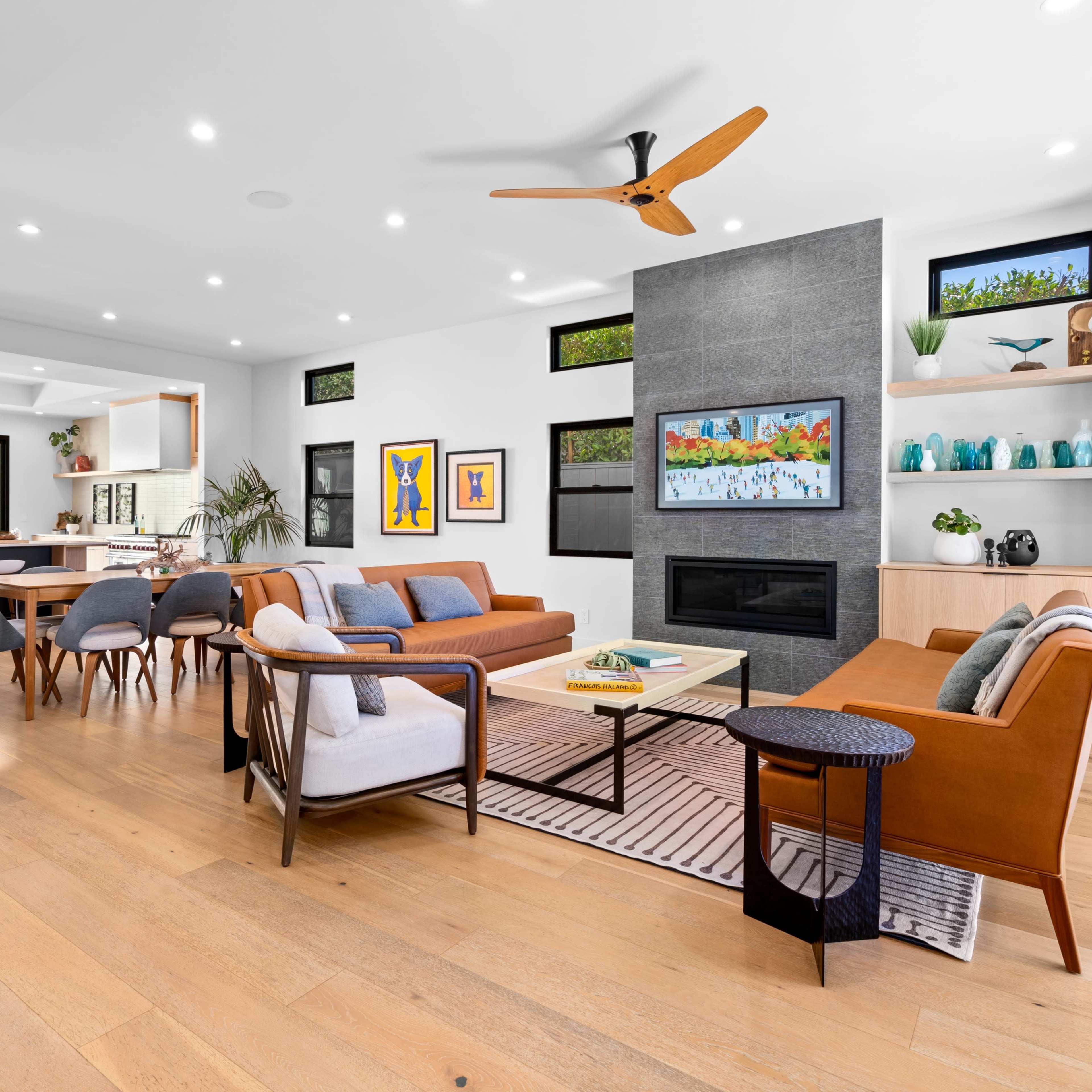 A modern, open-concept living area with a seating arrangement, a television, a kitchen bar, and large windows overlooking greenery.