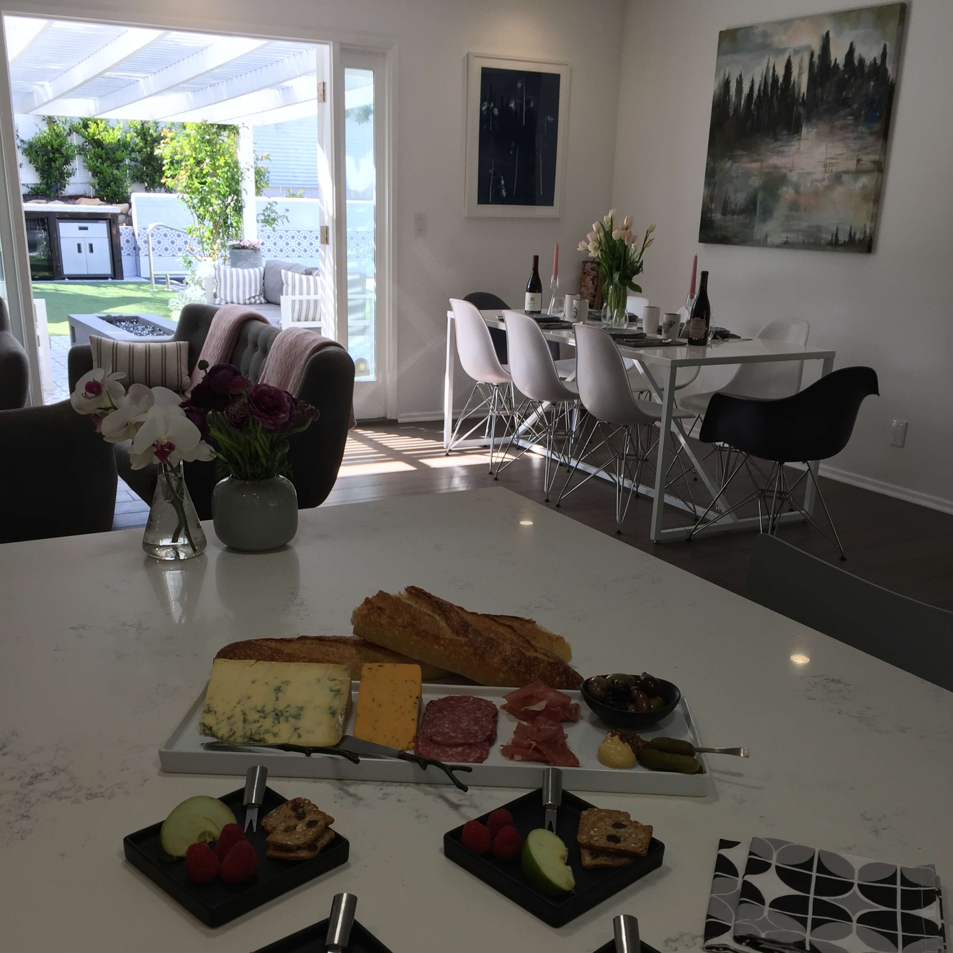 A kitchen counter is set with a charcuterie board, featuring assorted cheeses, meats, and fruits, while a dining area is visible in the background with a table set for a meal.